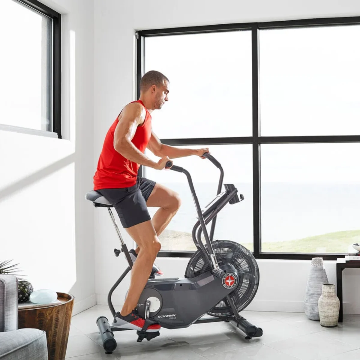 Homme en débardeur rouge monte un vélo à ventilateur gris à l'intérieur avec vue sur l'océan.