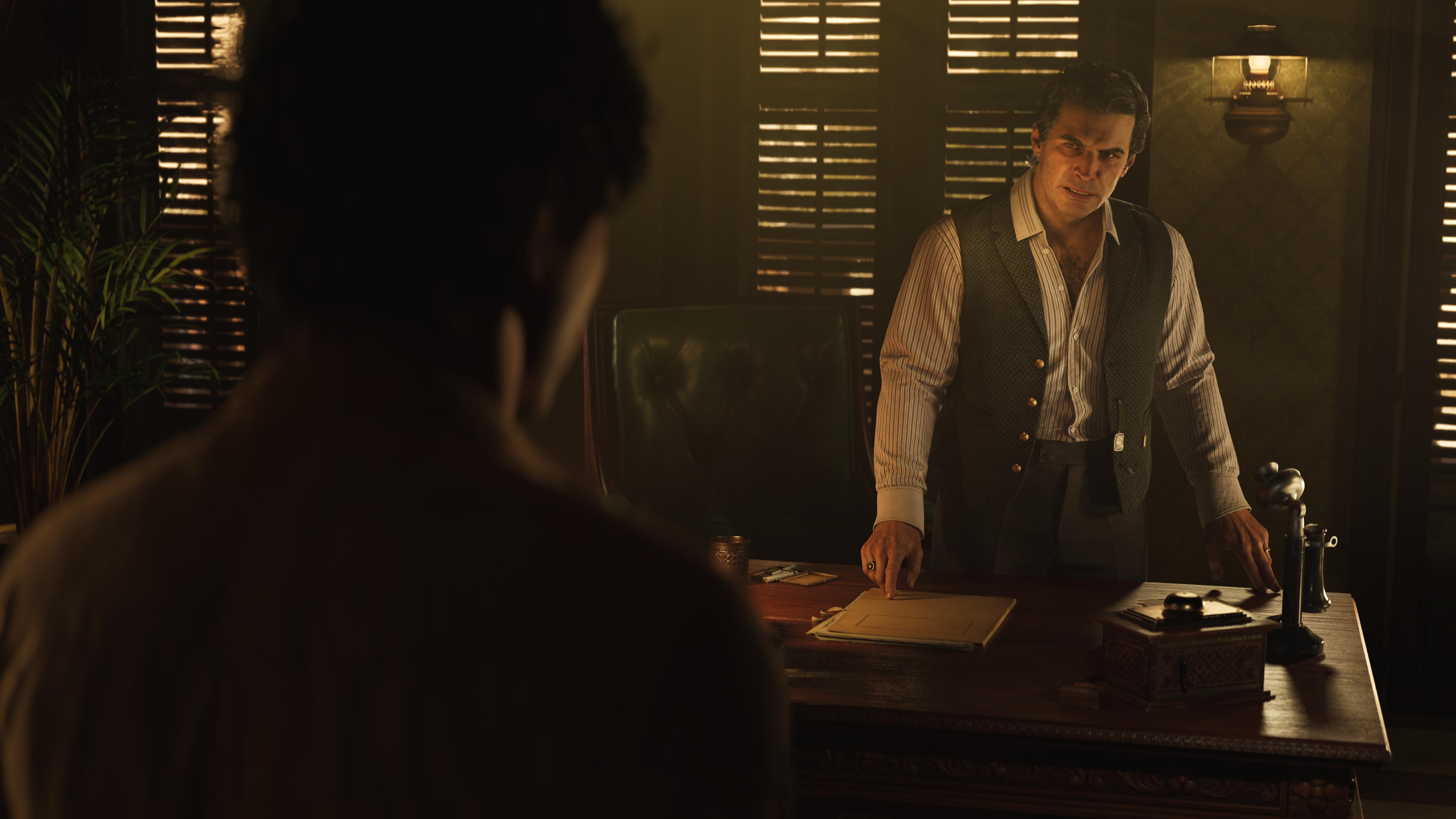 Mężczyzna w kamizelce i koszuli stoi za biurkiem. Druga osoba siedzi z przodu.