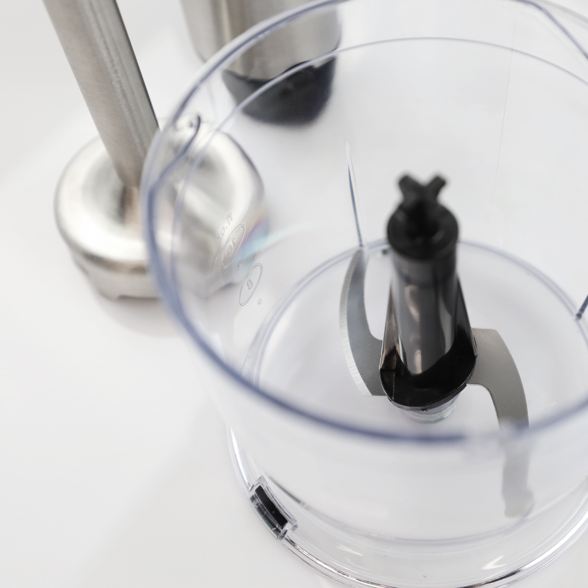 Zbliżenie przezroczystej misy malaksera z ostrzami i srebrną rączką blendera ręcznego.