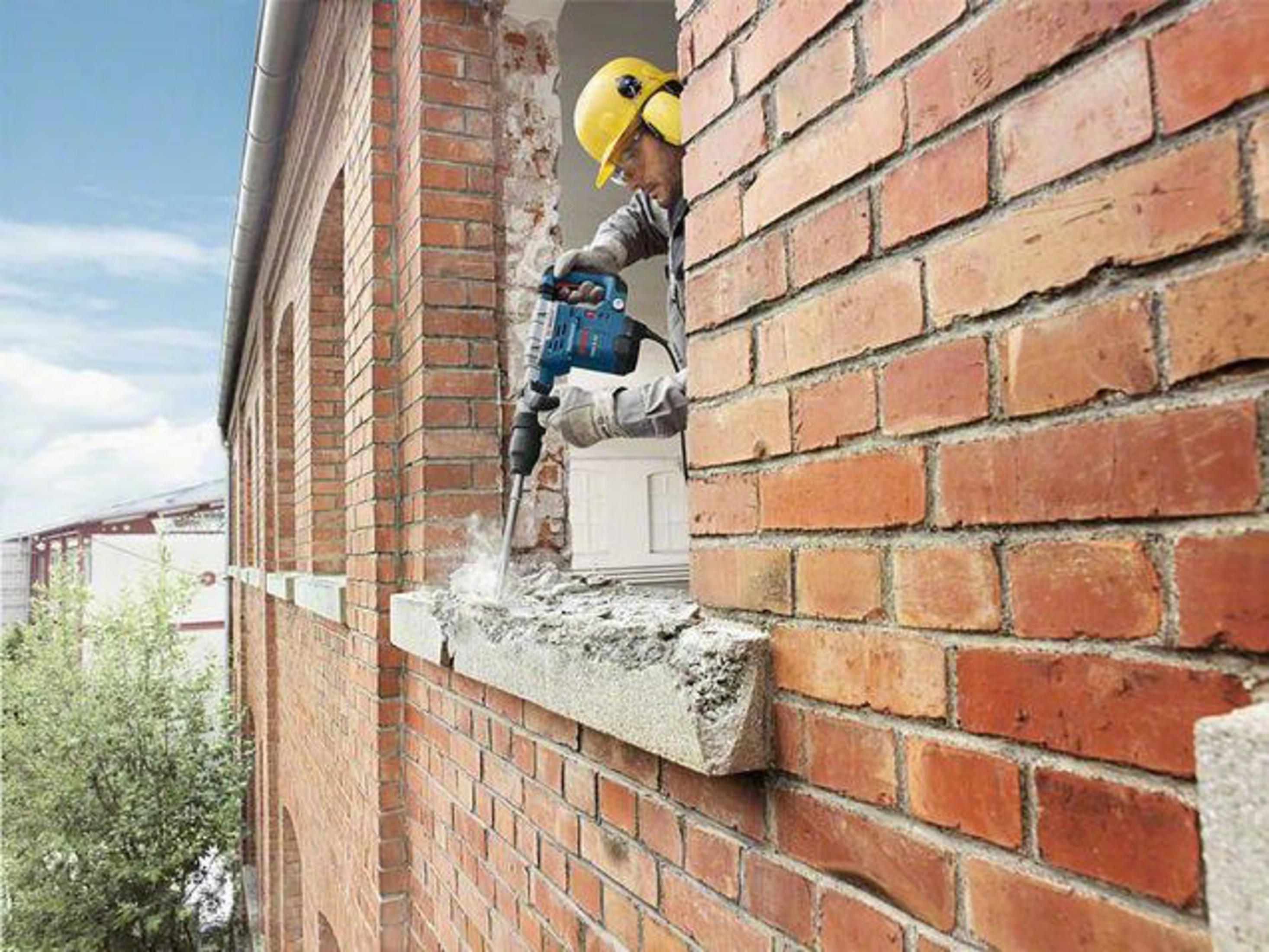 Hombre con casco amarillo usa martillo percutor en pared de ladrillo.