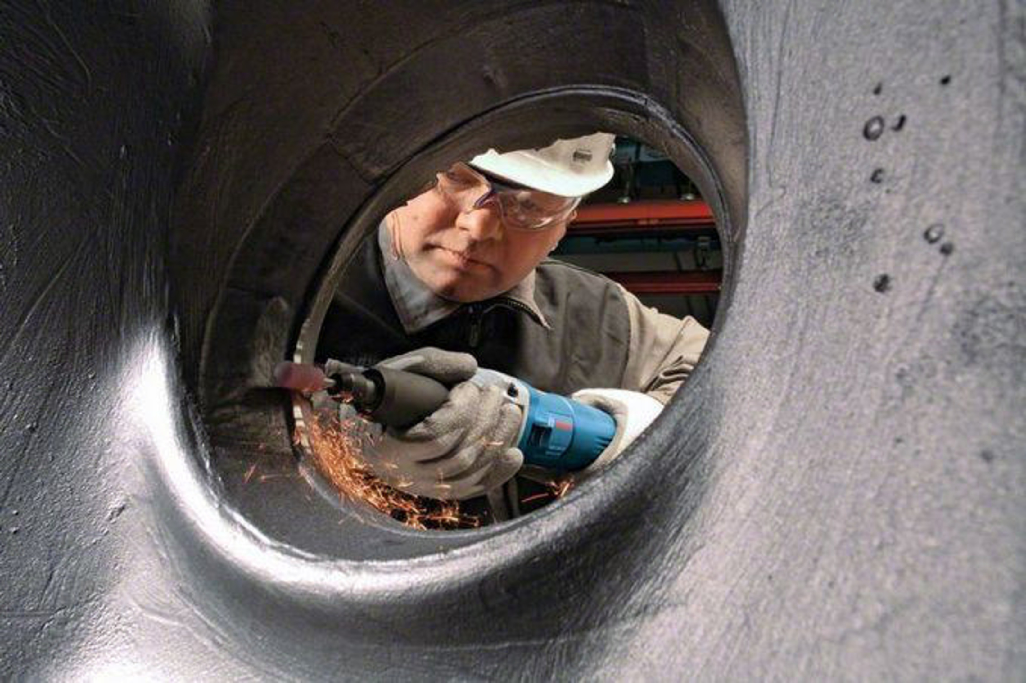 Hombre con casco y gafas afilando borde metálico con herramienta, creando chispas.
