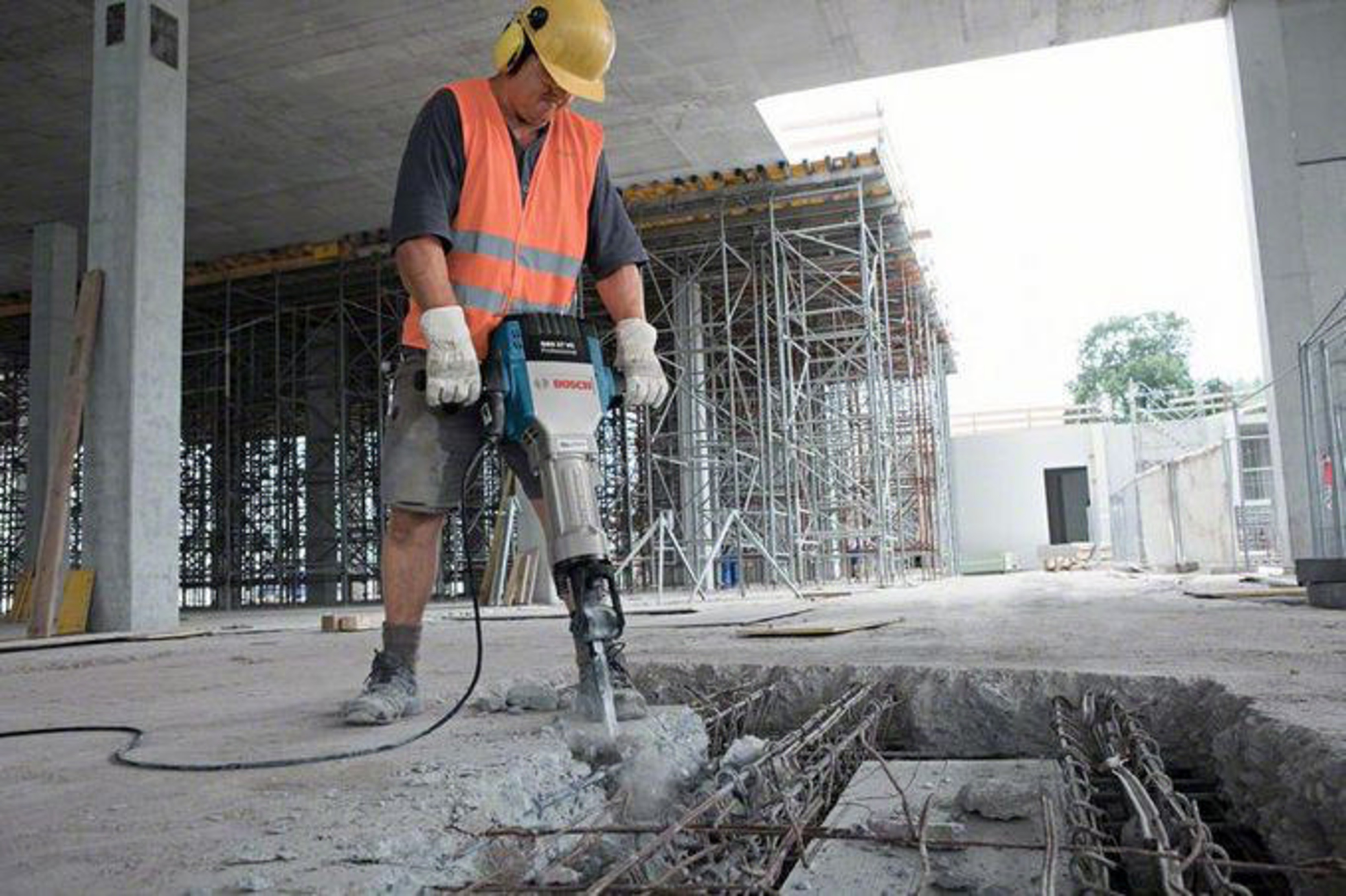 Adam kırıcı çekiçle inşaatta beton kırıyor, arkada iskele var.