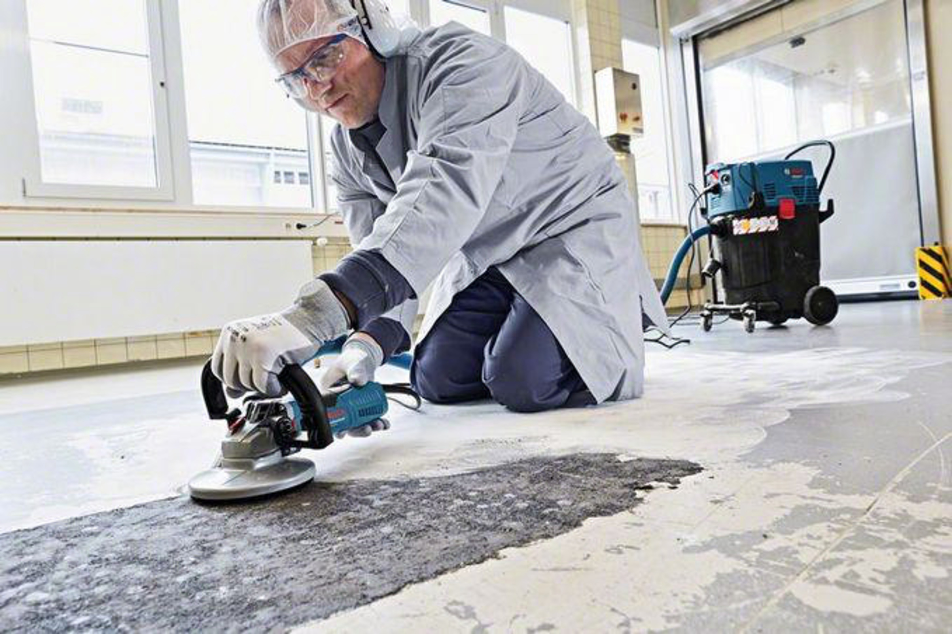 Mann im Laborkittel schleift Betonboden mit Elektrowerkzeug. Staubsauger daneben.