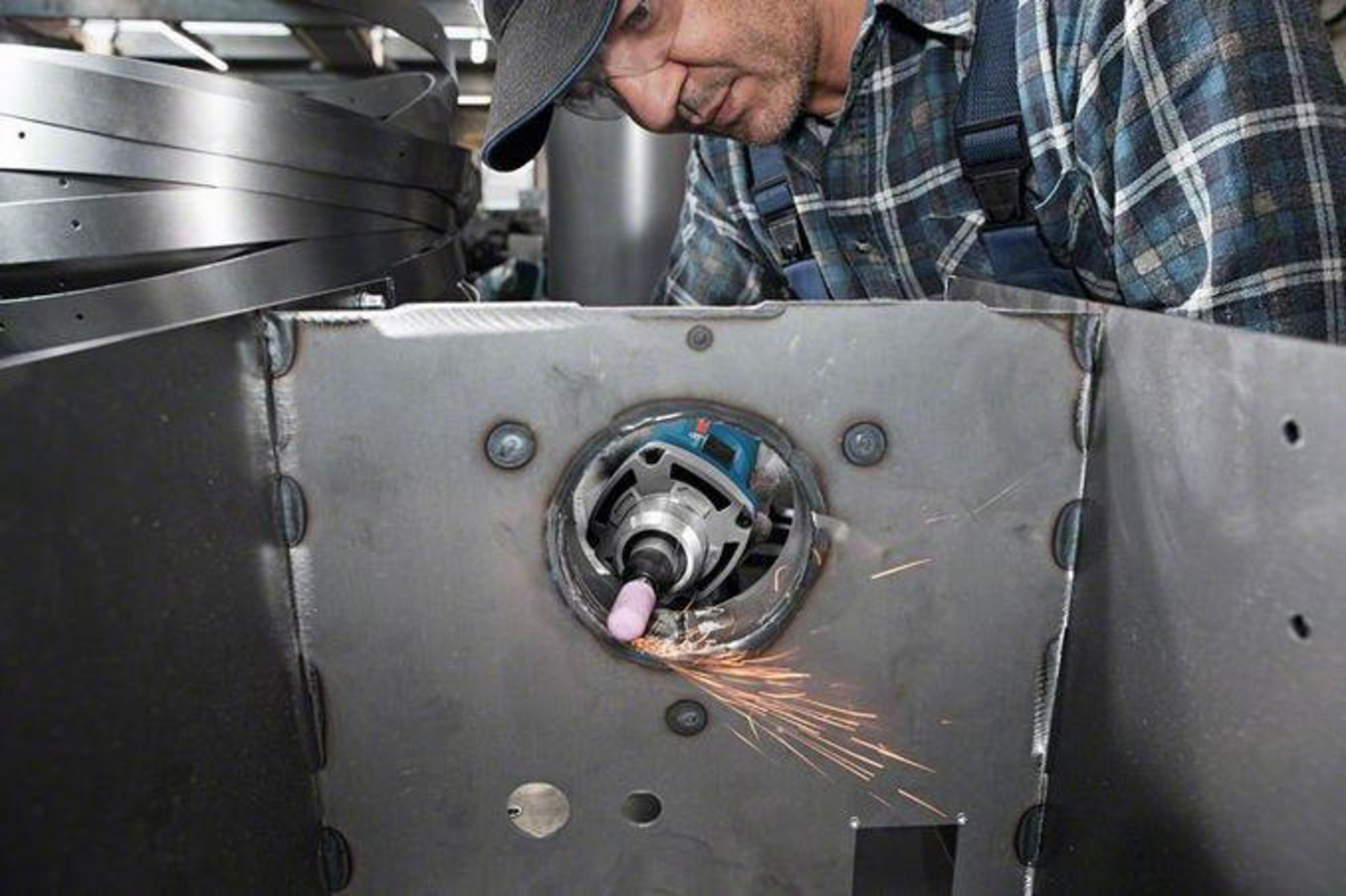 Un hombre con ropa de trabajo a cuadros azules lija metal con una herramienta.