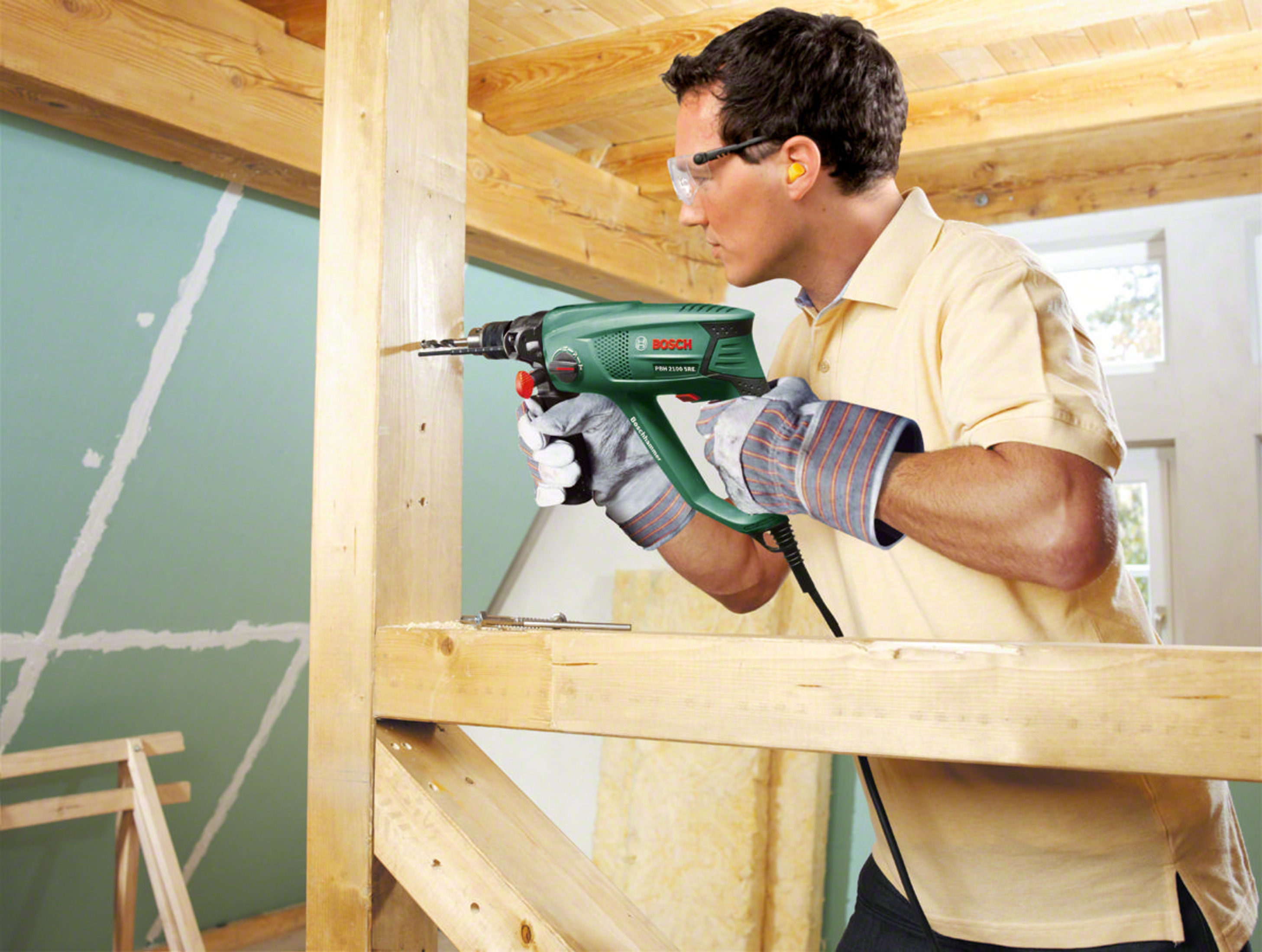 Uomo fora legno con trapano Bosch verde, indossa occhiali e guanti.