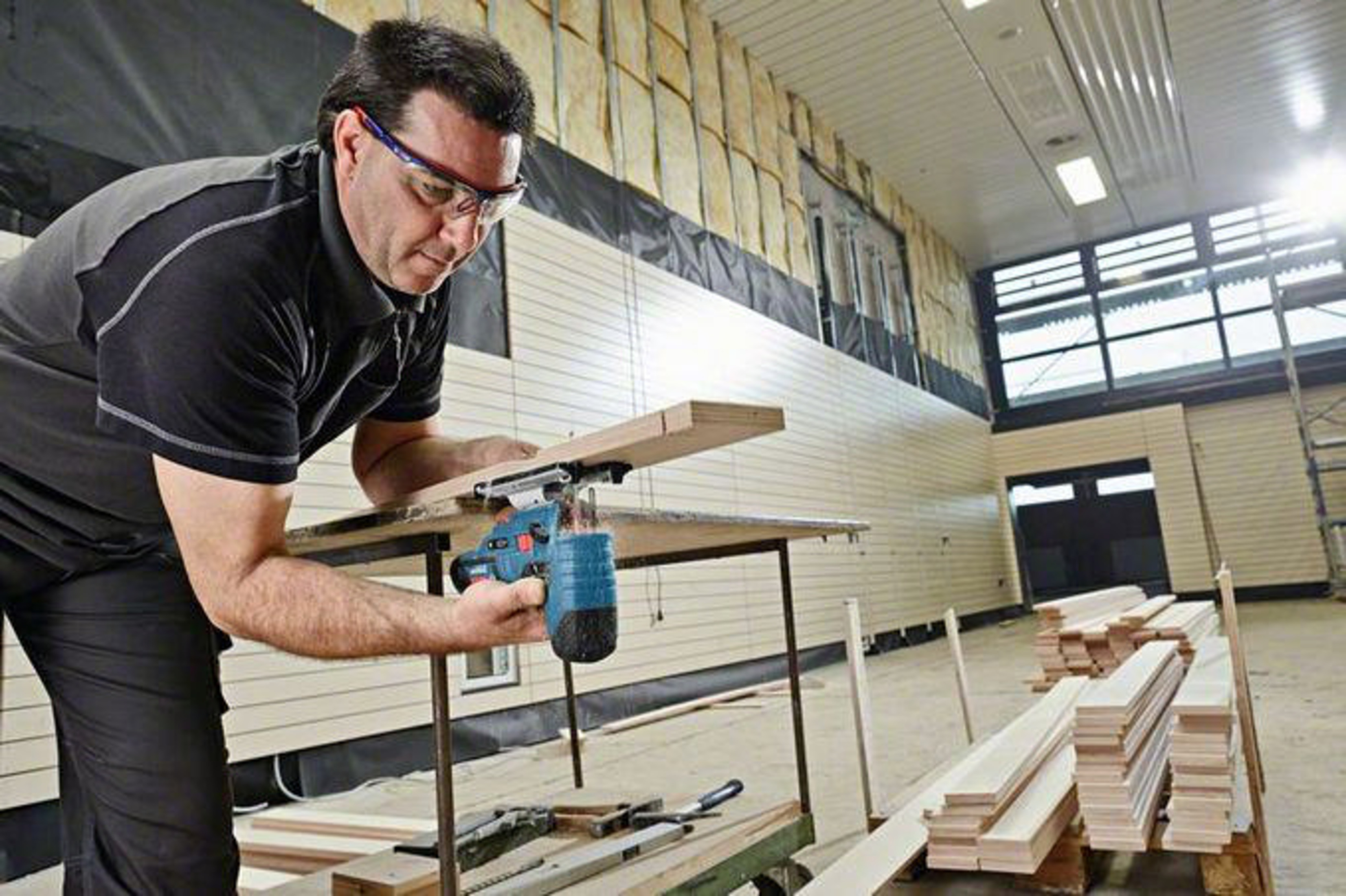 Ein Mann mit Schutzbrille schneidet mit einer Stichsäge ein Holzbrett auf einer Werkbank auf einer Baustelle.