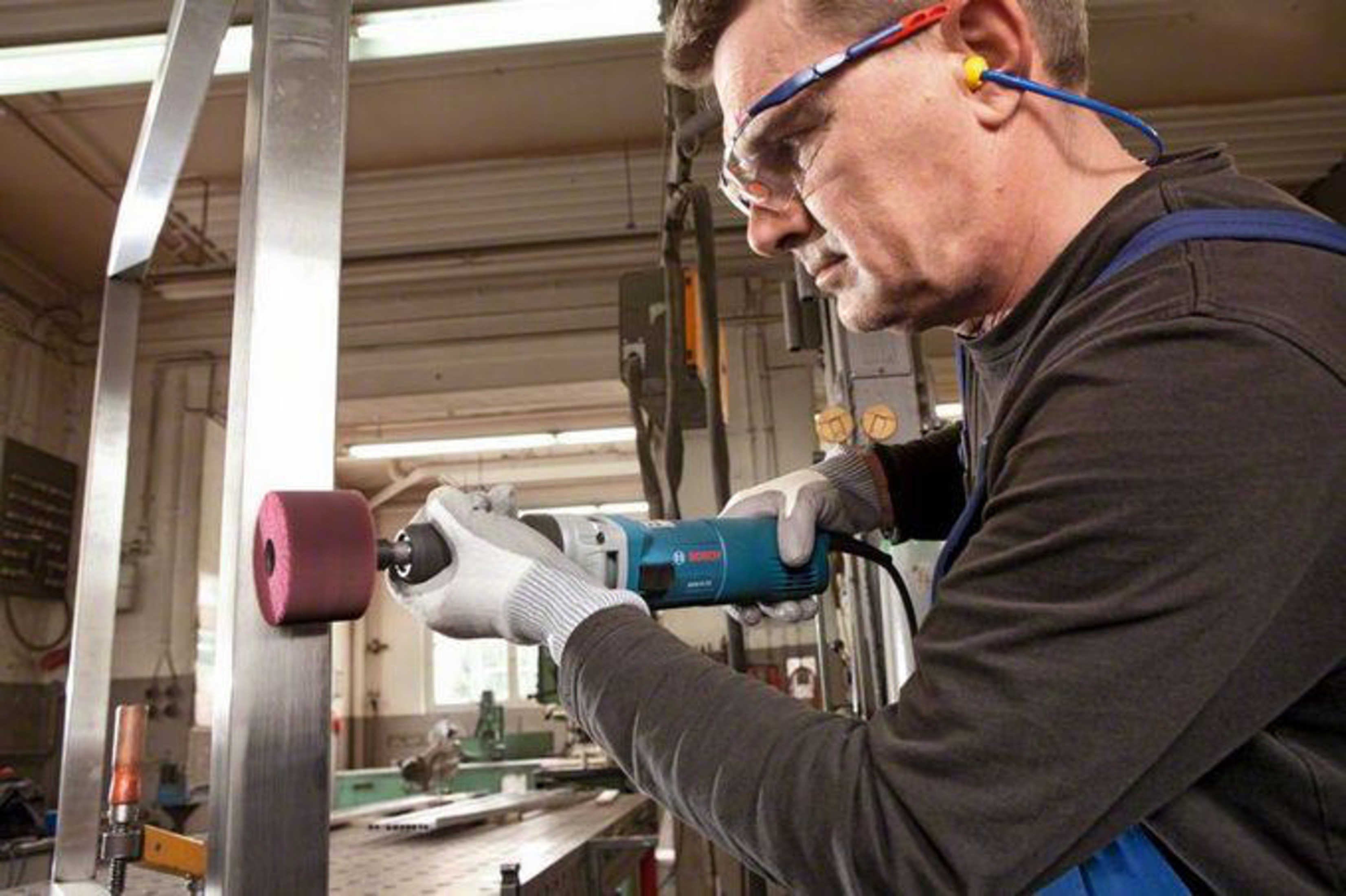 Mężczyzna w okularach ochronnych i zatyczkach do uszu używa niebieskiego elektronarzędzia z czerwoną tarczą ścierną na metalowej belce.