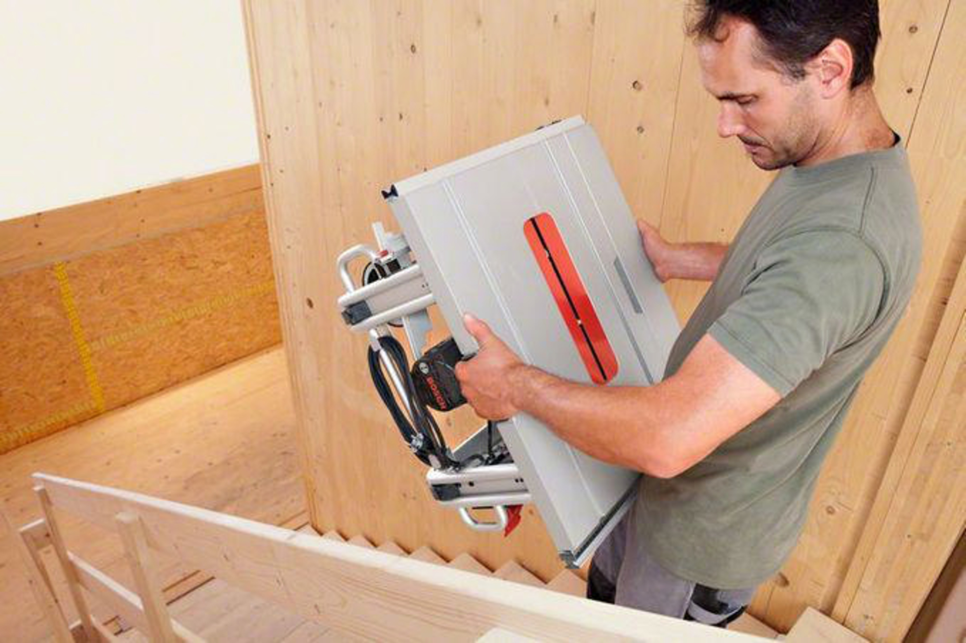 Hombre sube sierra de mesa por escalera de madera, pared de madera detrás.