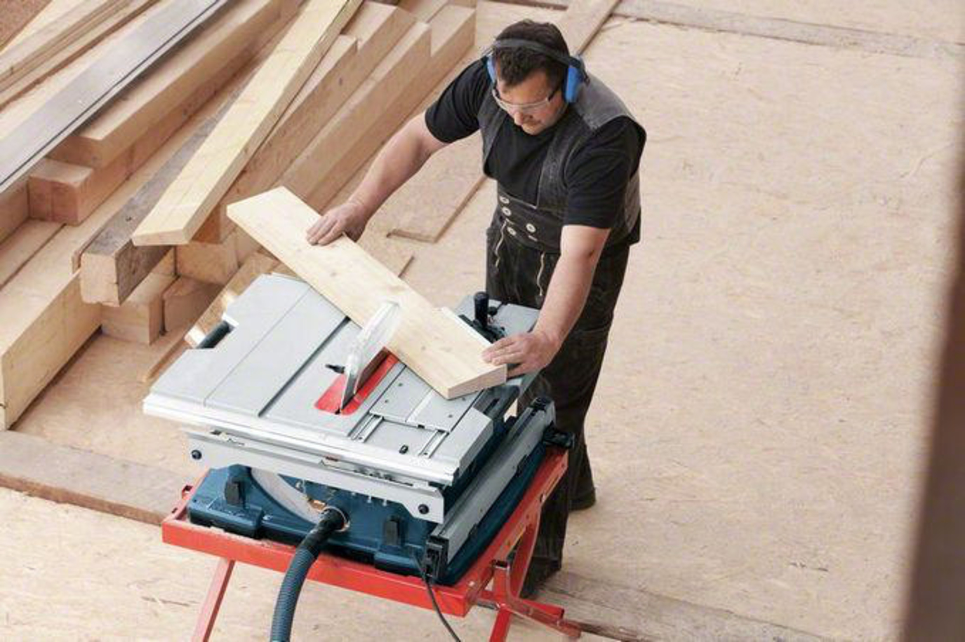 Uomo taglia legno su banco sega. Molte assi di legno sparse intorno.