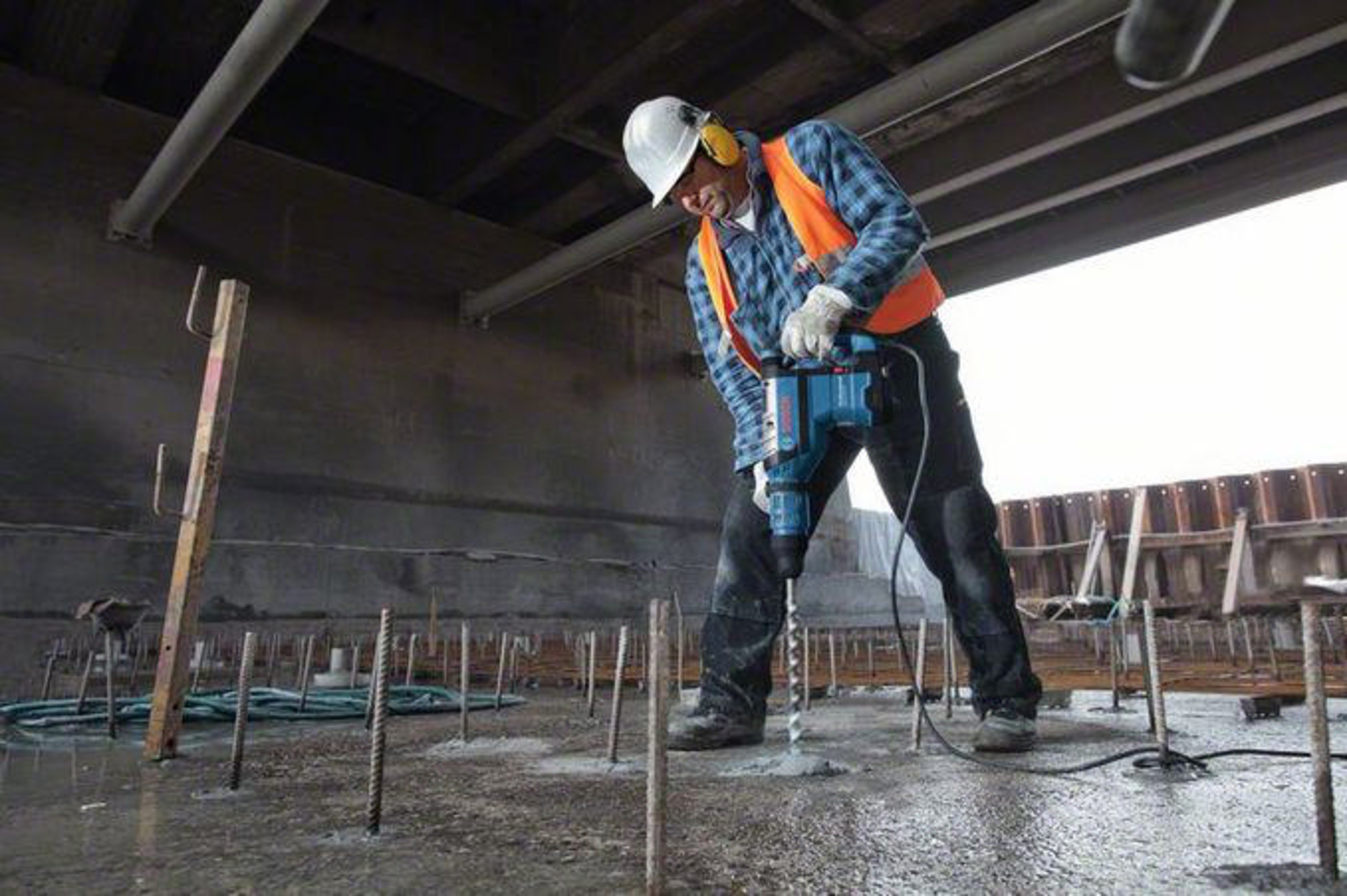 Mann bohrt in Beton, trägt Helm, Weste und Gehörschutz.