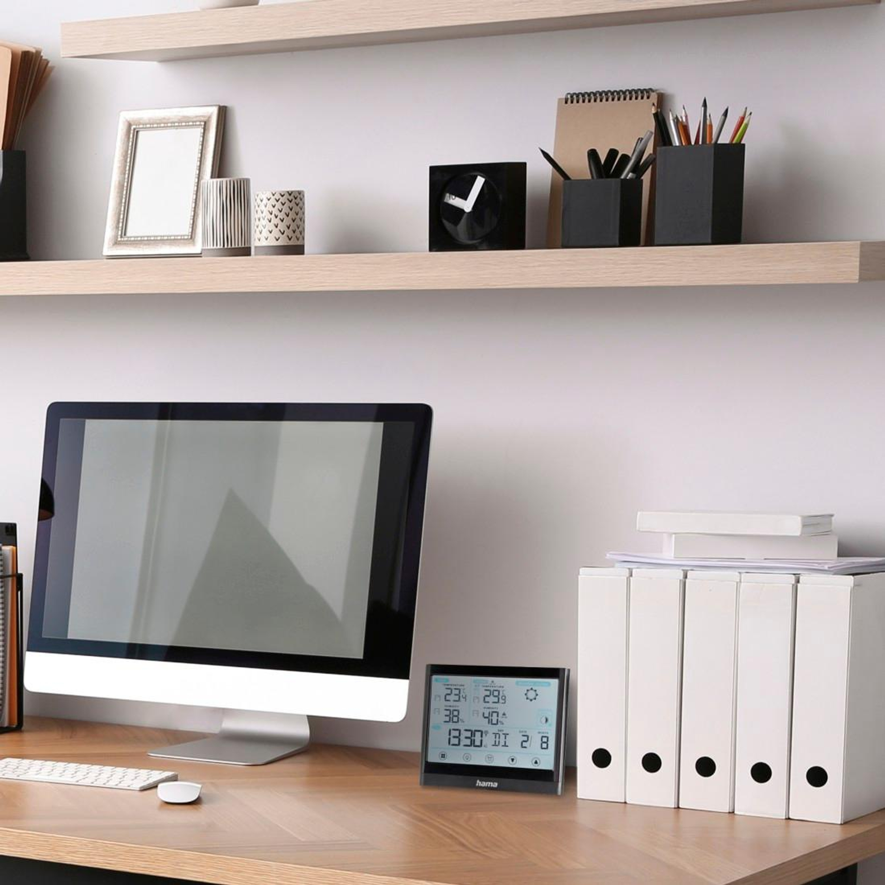 Drewniane biurko z komputerem, białymi segregatorami i cyfrową stacją pogodową. Dwie jasne drewniane półki z dekoracjami.