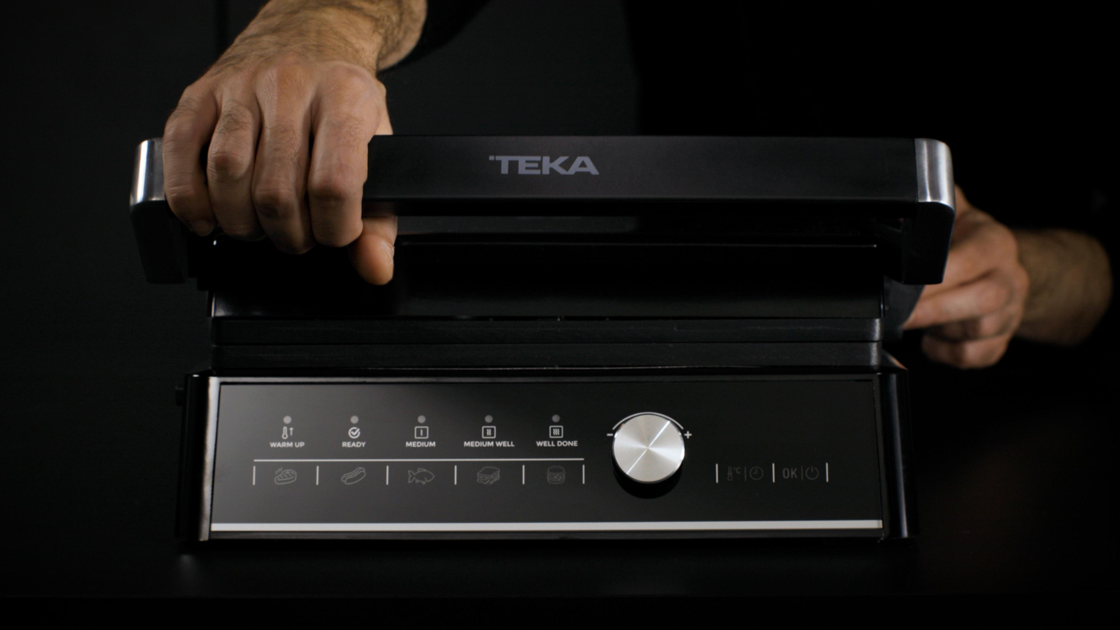 Una persona abre una parrilla negra con logo TEKA y controles digitales.