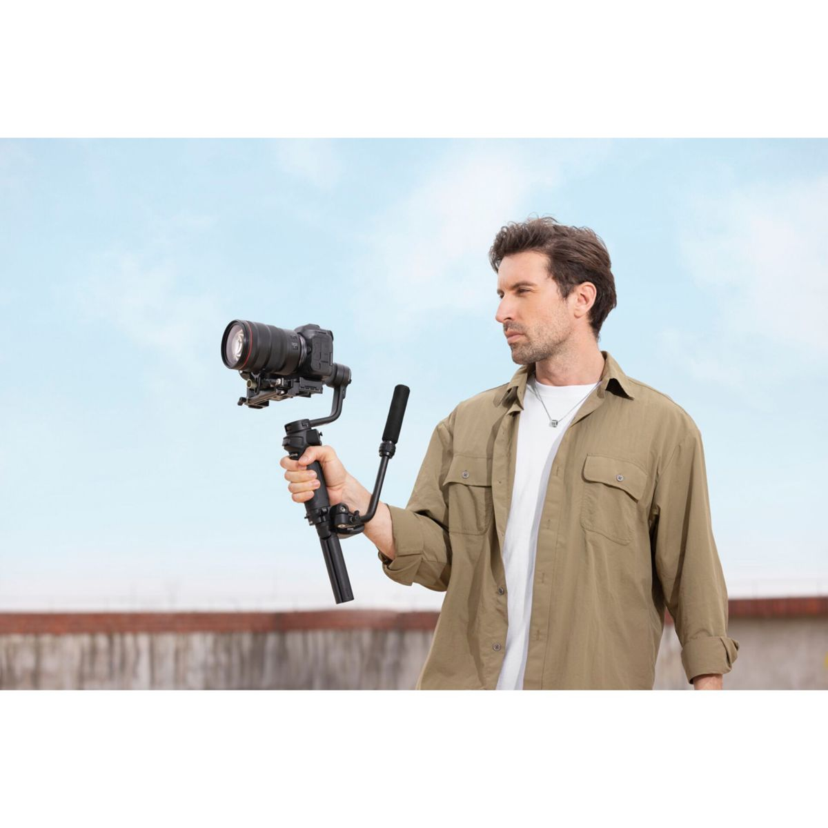 Ein Mann mit einem Gimbal und einer Kamera in der Hand steht vor blauem Himmel.