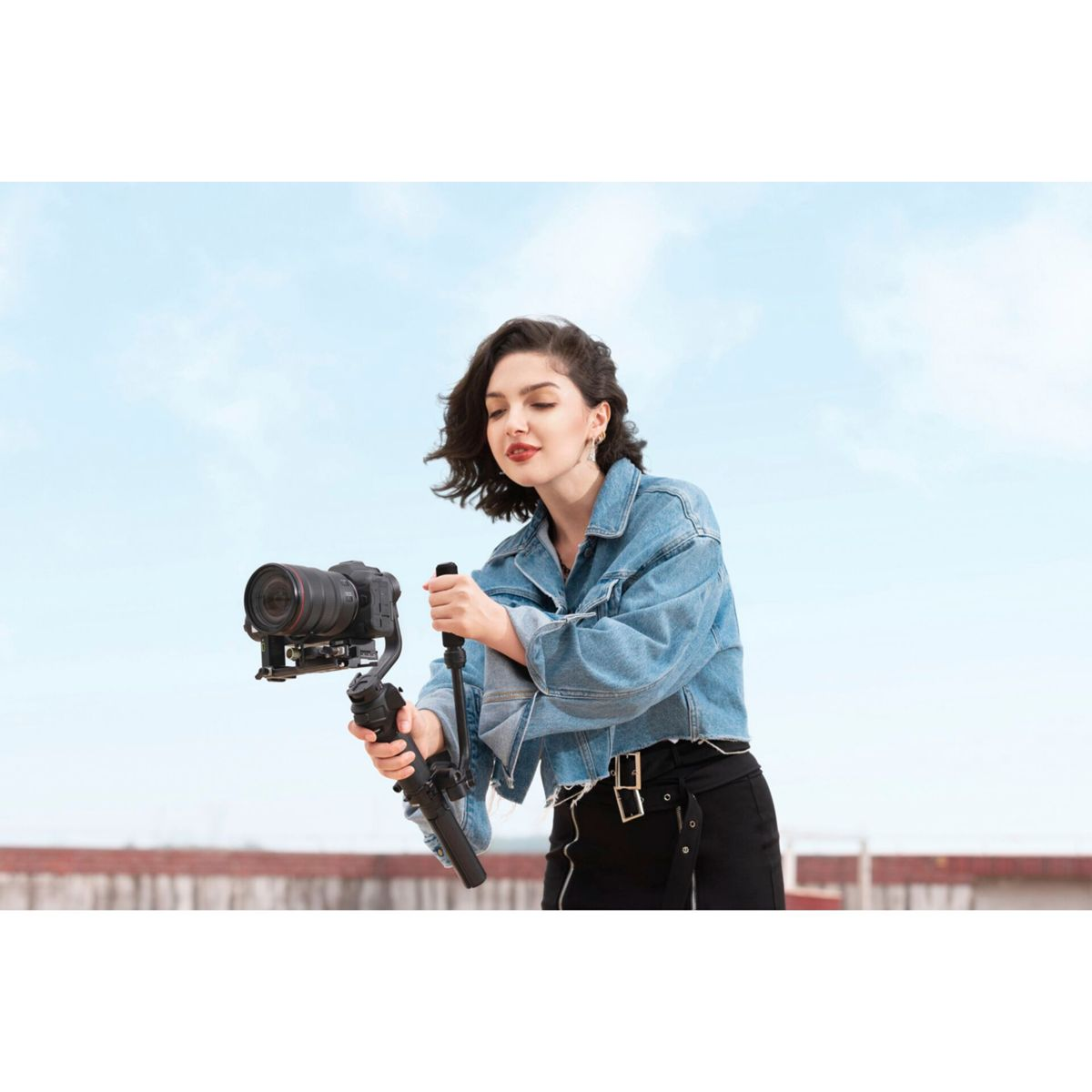 Eine Frau in Jeansjacke hält eine Kamera mit Gimbal vor blauem Himmel.