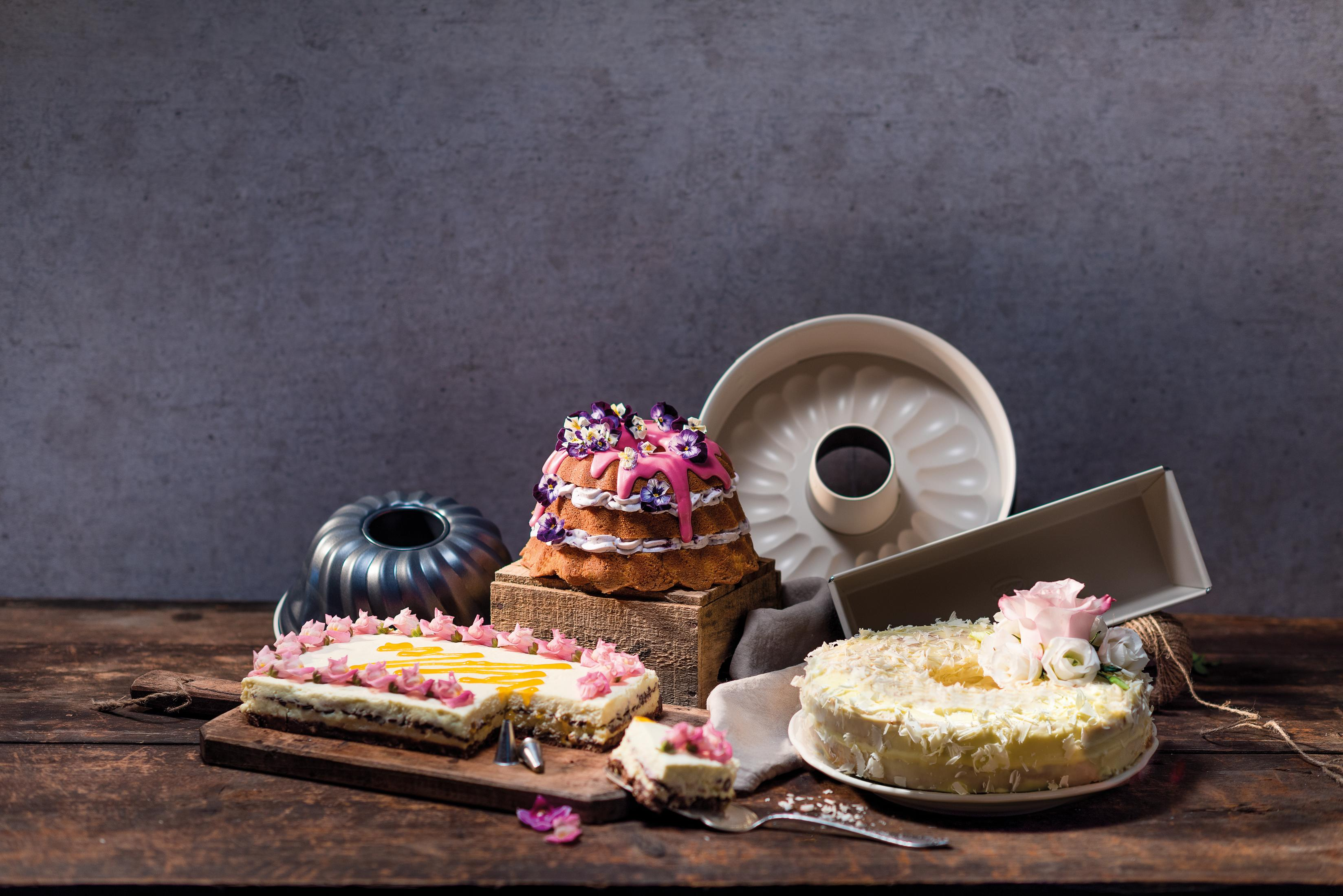Drei Kuchen auf einem Holztisch. Ein rosa glasierter Kuchen mit lila Blüten, ein rechteckiger Kuchen mit gelben und rosa Akzenten und ein weißer Kuchen mit weißen Schokoladenraspeln.
