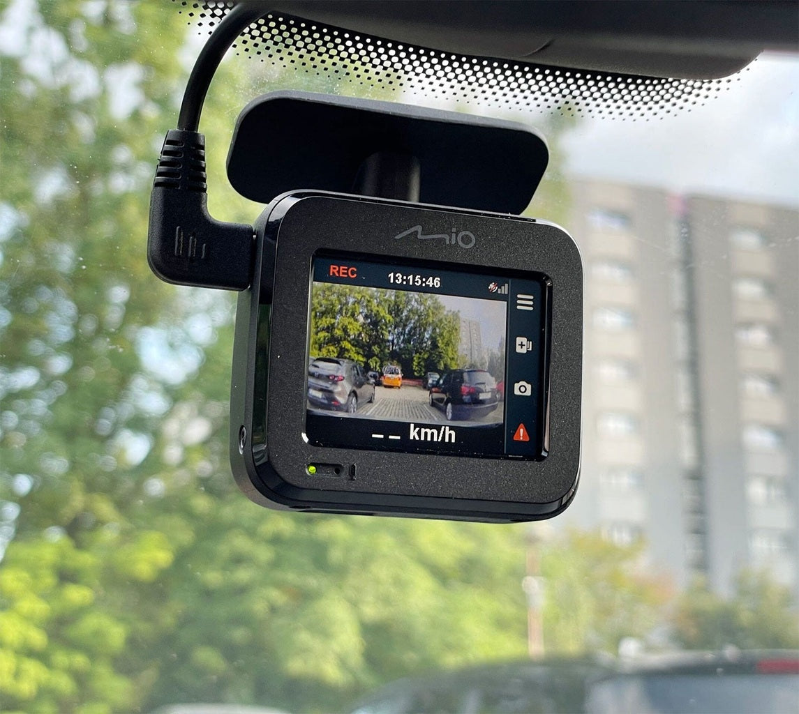 Armaturenbrettkamera zeigt Autos auf einem Parkplatz mit Bäumen und Gebäuden im Hintergrund.