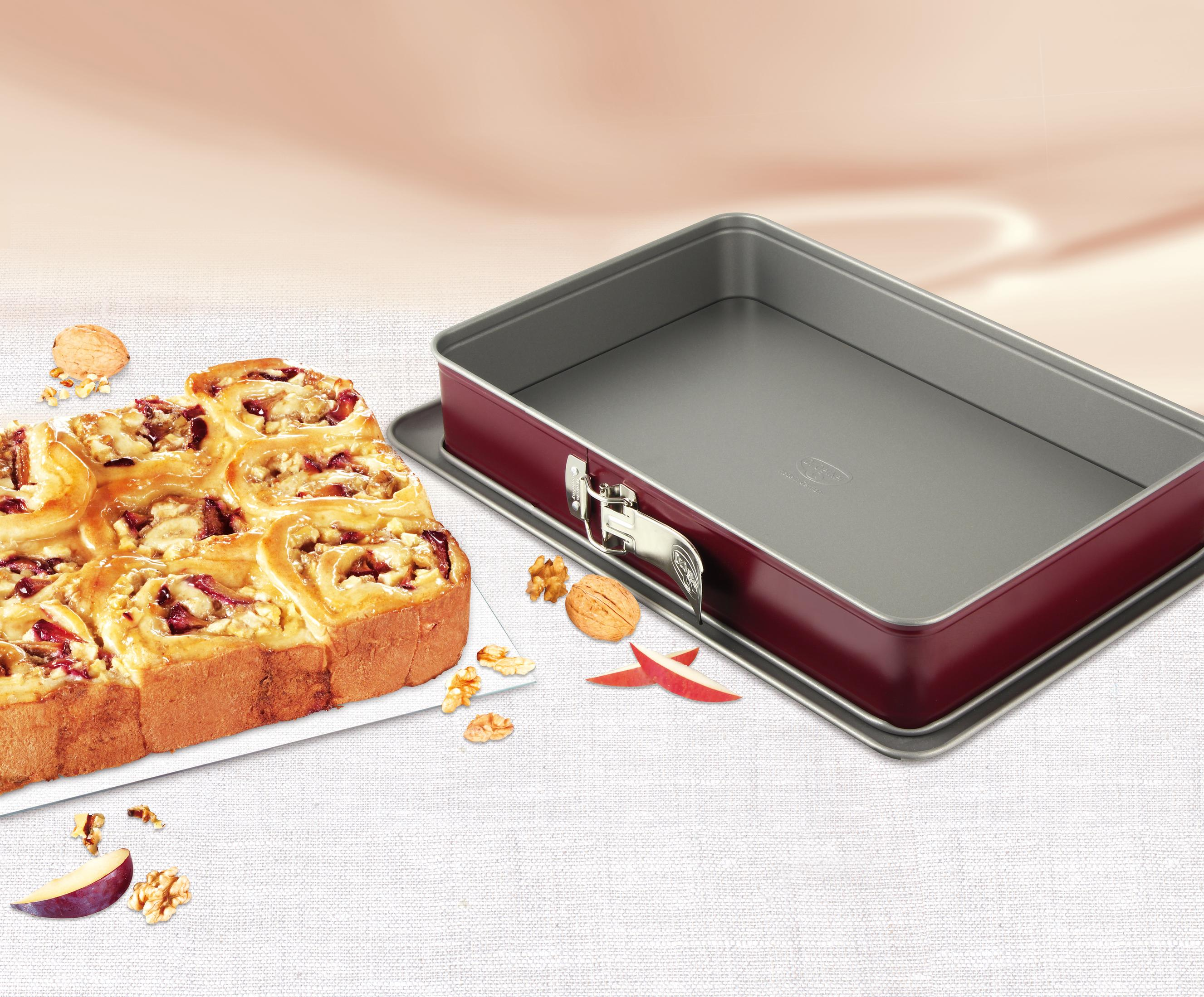 Ein rechteckiger Backblech mit Burgunderroten Seiten neben einem Kuchen mit Nüssen und Früchten.
