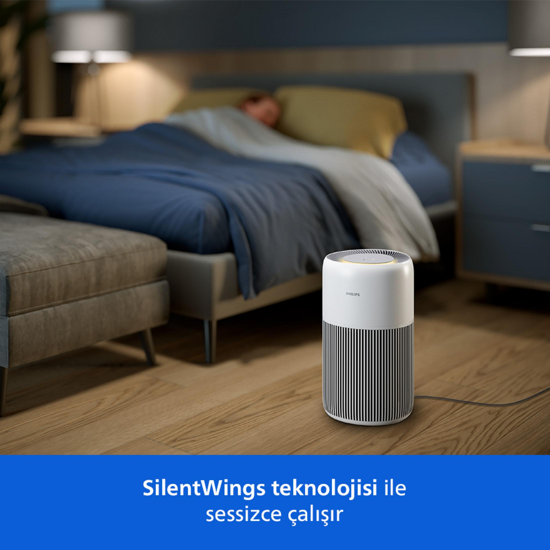 Biały oczyszczacz powietrza Philips stoi na drewnianej podłodze obok łóżka, na którym śpi osoba.