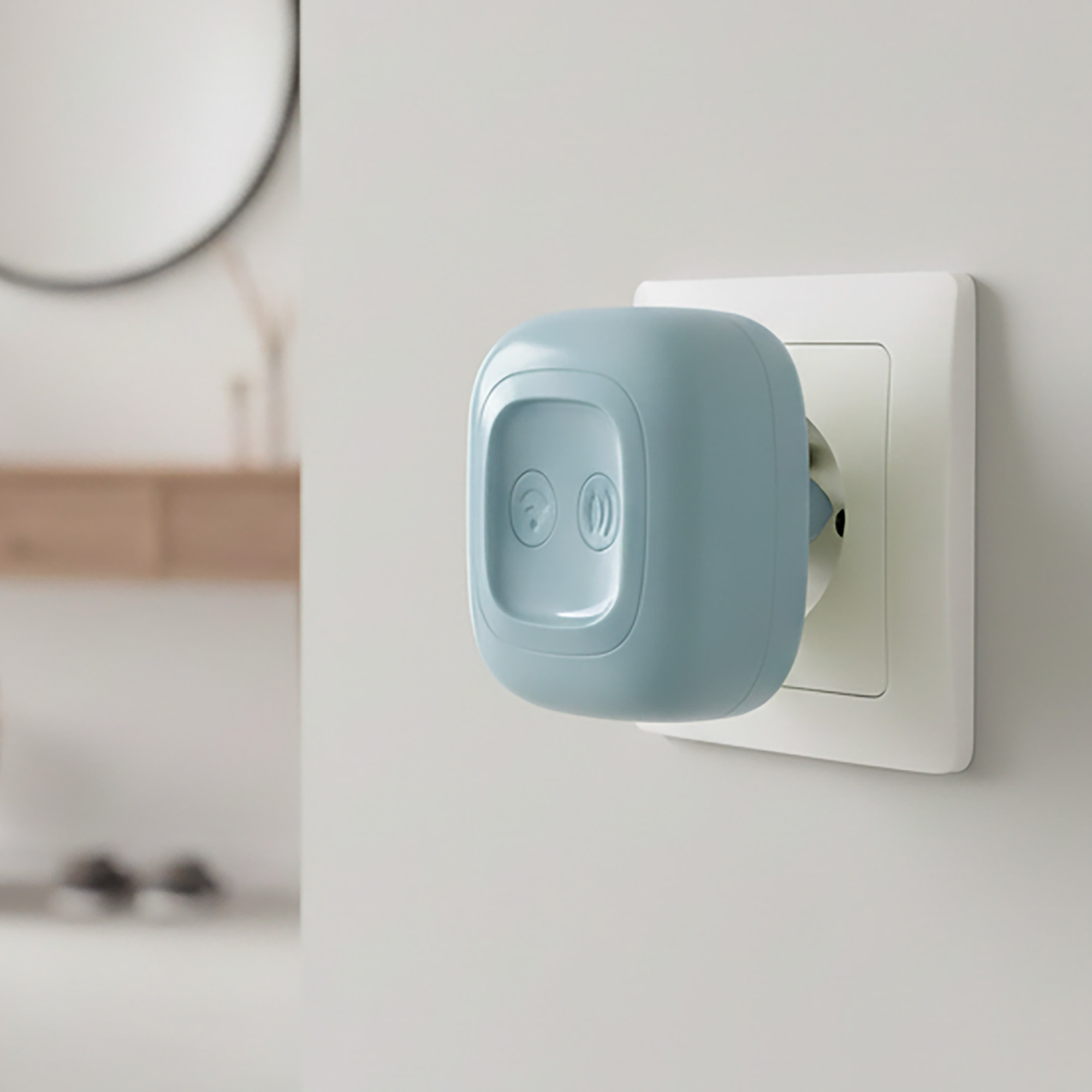 Un dispositivo azul claro está enchufado en un enchufe de pared blanco. Un espejo está desenfocado a la izquierda.