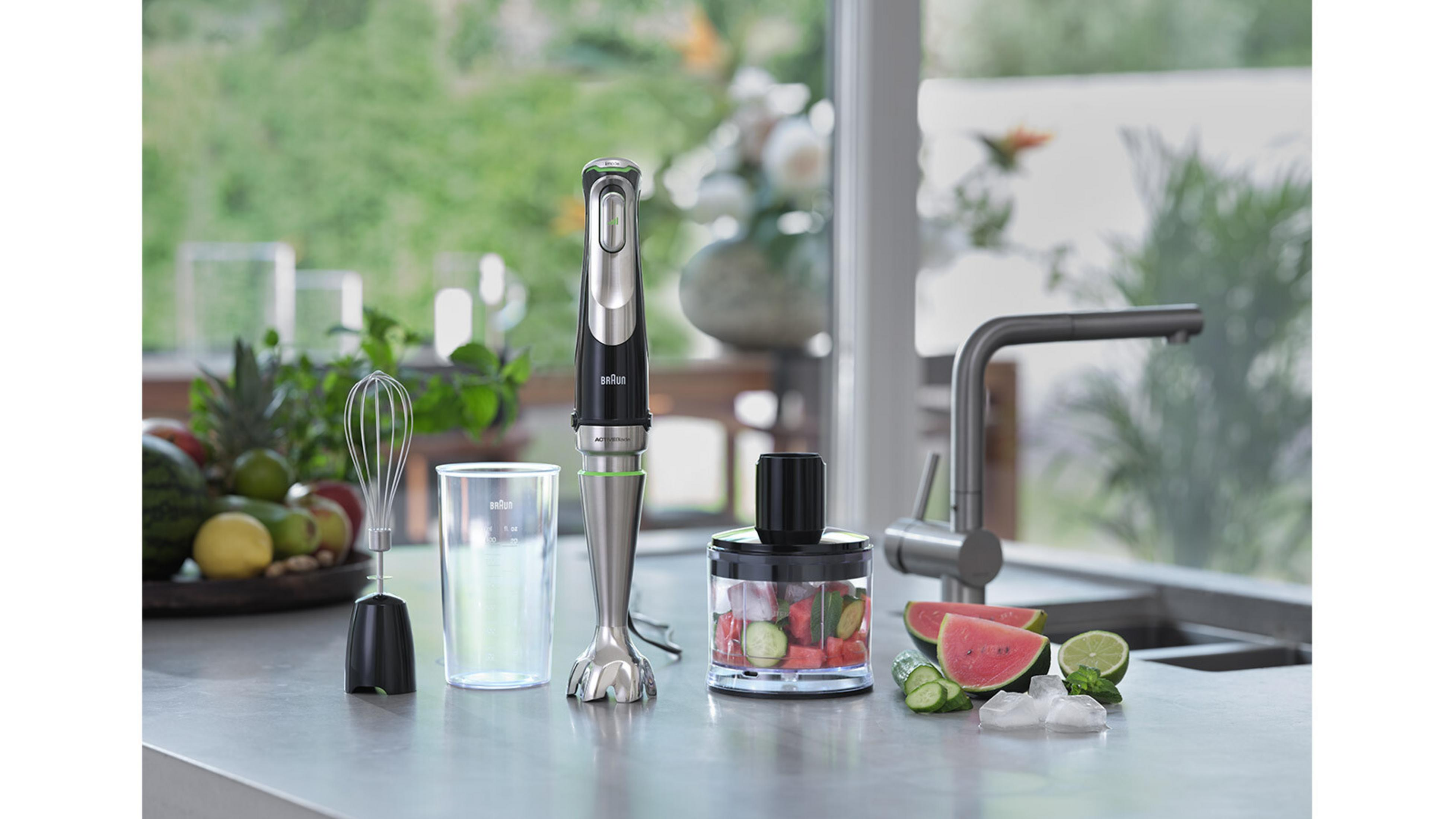 Een staafmixer met accessoires, watermeloen, komkommer en ijsblokjes op een aanrecht.