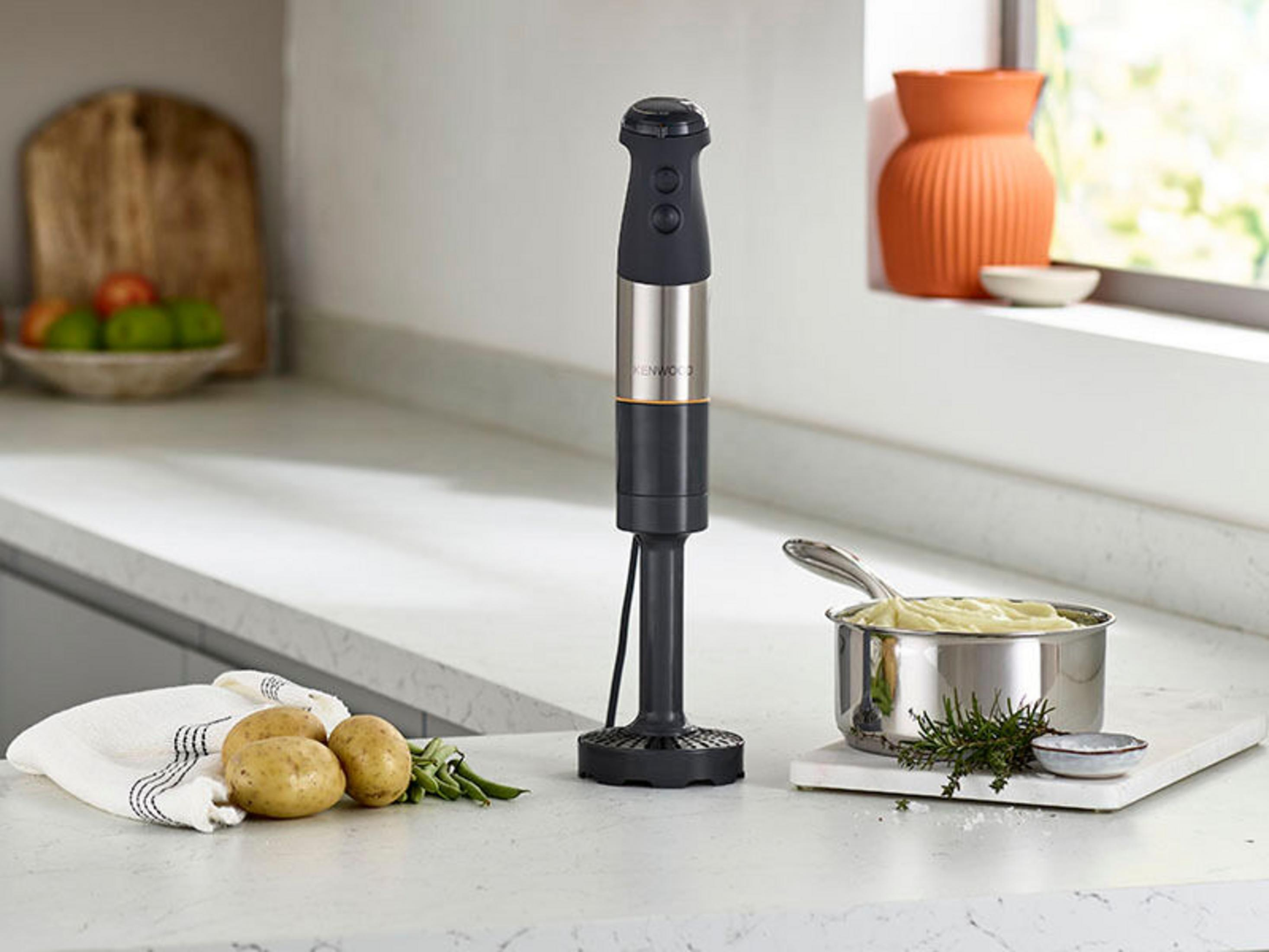 Blender ręczny stoi na blacie obok ziemniaków i garnka z puree ziemniaczanym.