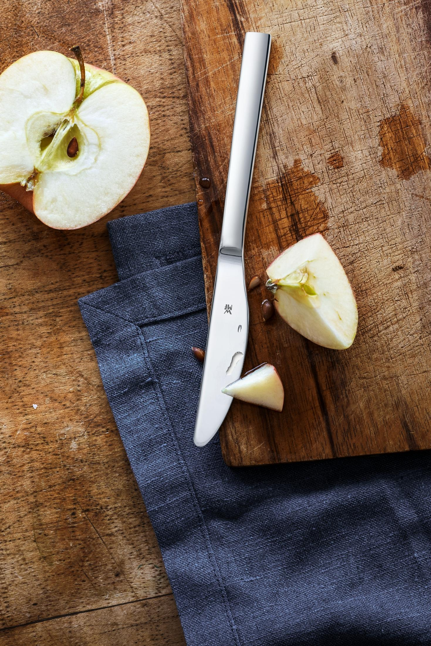 Ein halber Apfel, ein Messer und Apfelstücke auf einem Holzbrett.