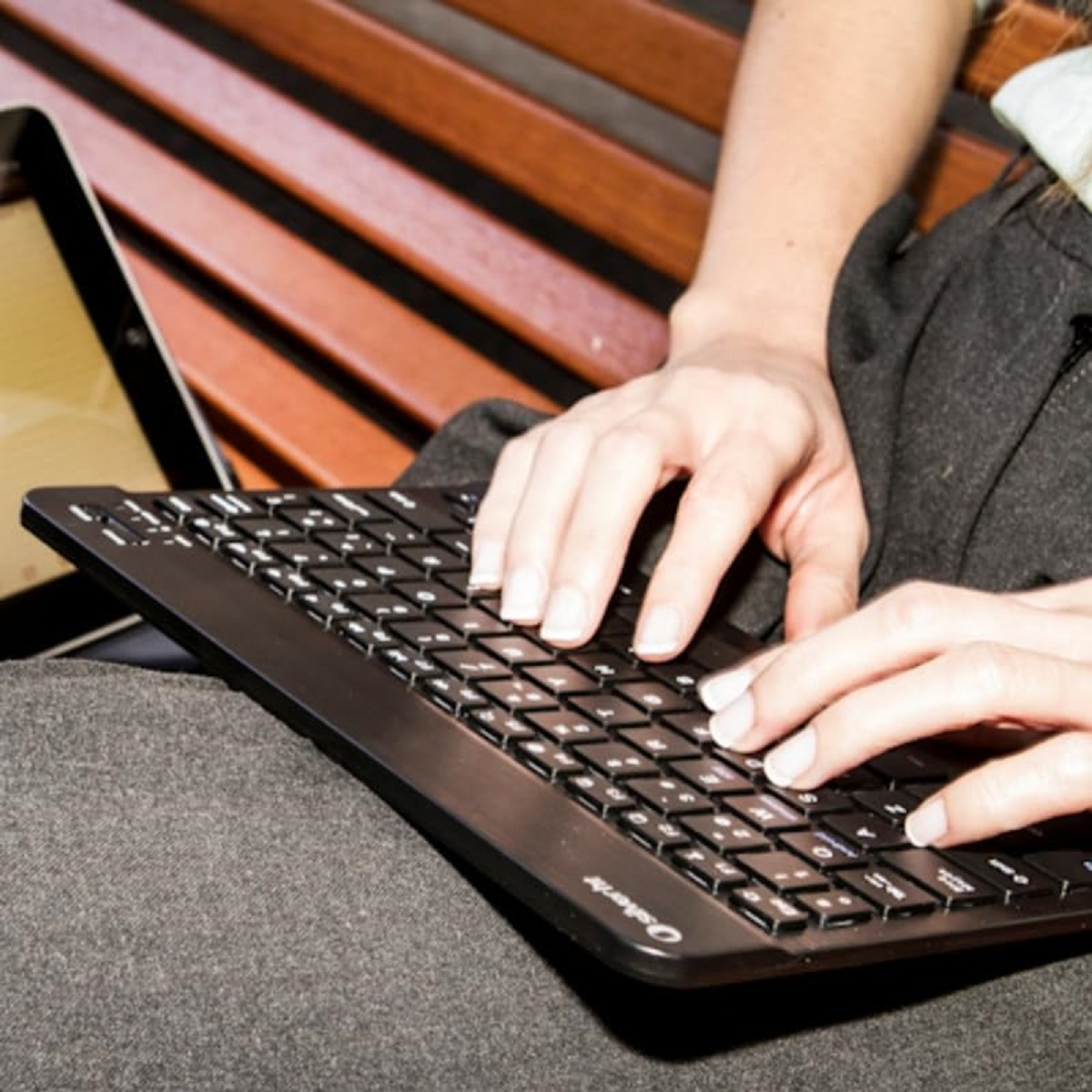 Primer plano de las manos de una persona escribiendo en un teclado inalámbrico negro, con una tableta y un banco de madera detrás.