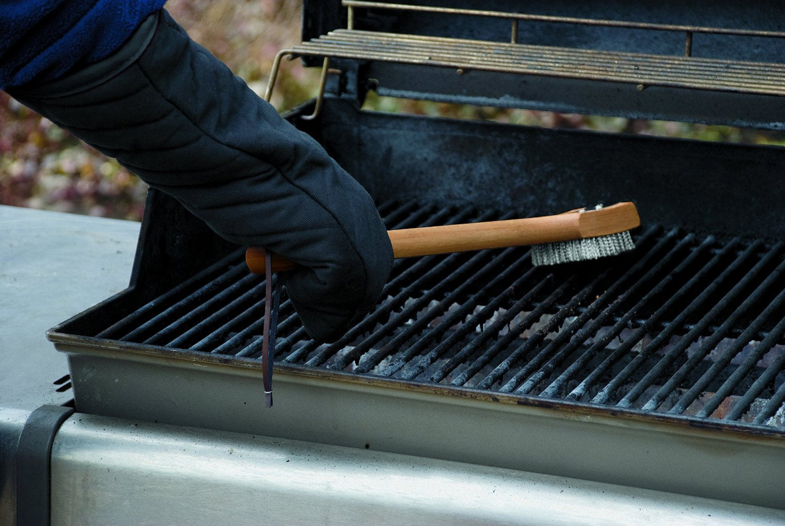 Eine Hand in einem schwarzen Handschuh reinigt den Grillrost mit einer Bürste.