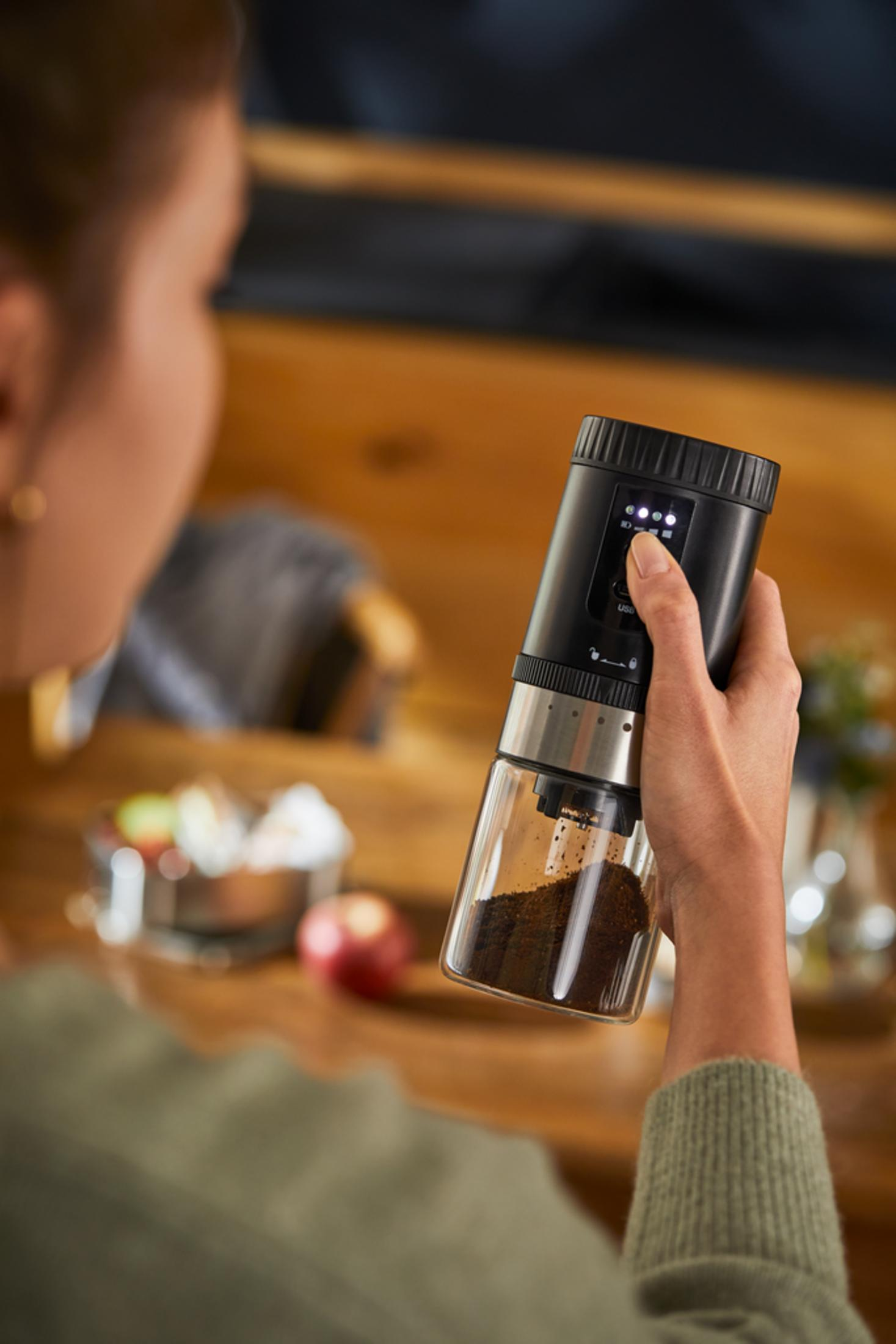 Eine Hand hält eine schwarze elektrische Kaffeemühle mit Glasboden, gefüllt mit Kaffeepulver.