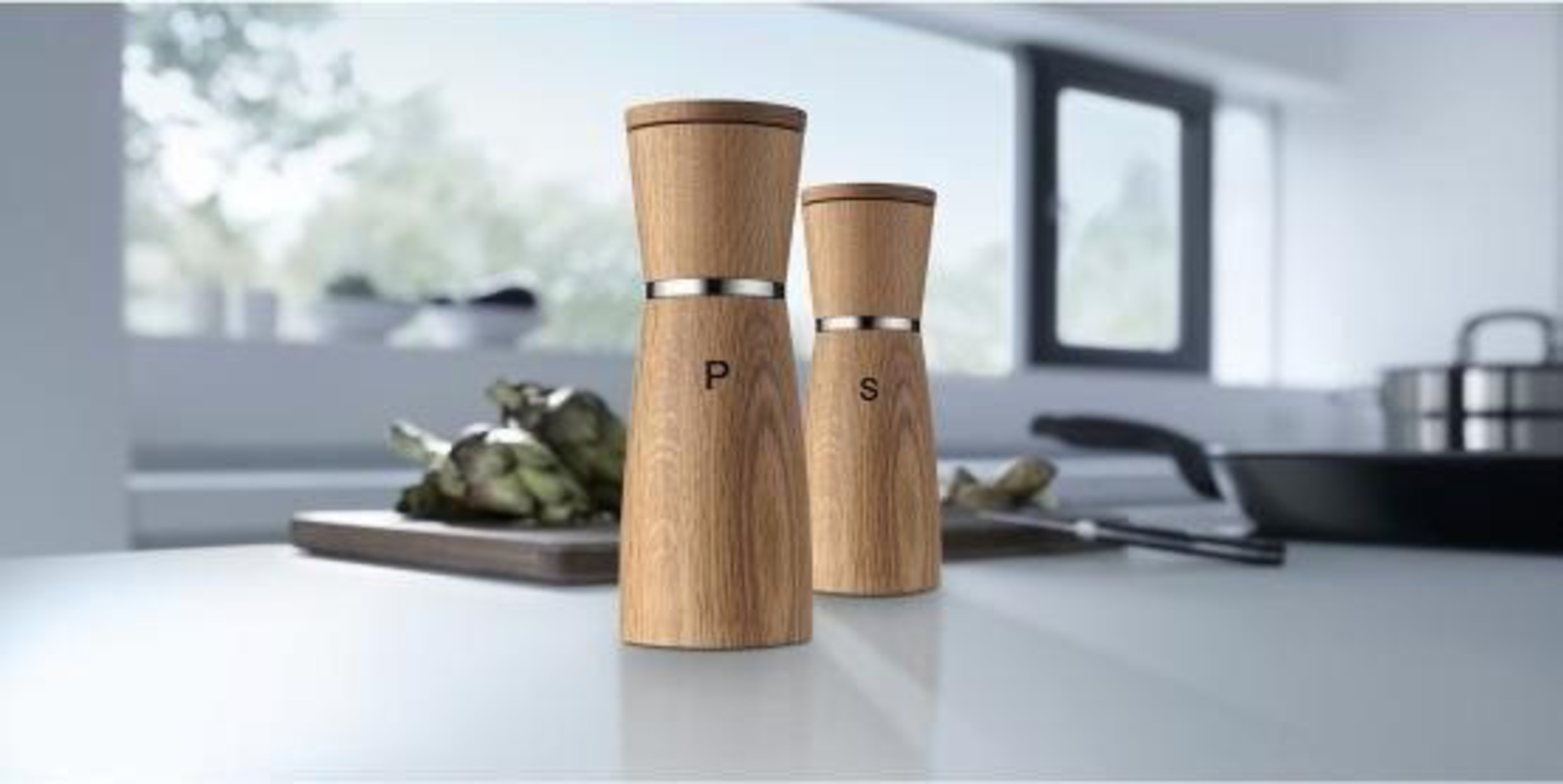 Zwei Holz-Salz-und-Pfeffermühlen auf einer Arbeitsplatte, mit Artischocken und Pfanne.