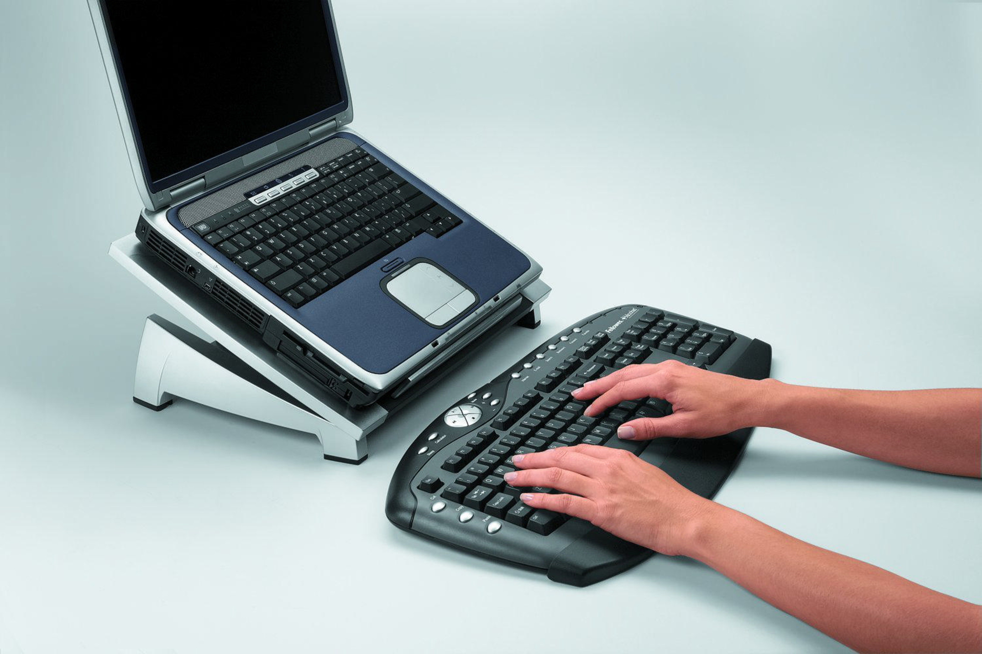 Ein Laptop auf einem Ständer neben einer Tastatur, auf der Hände tippen.