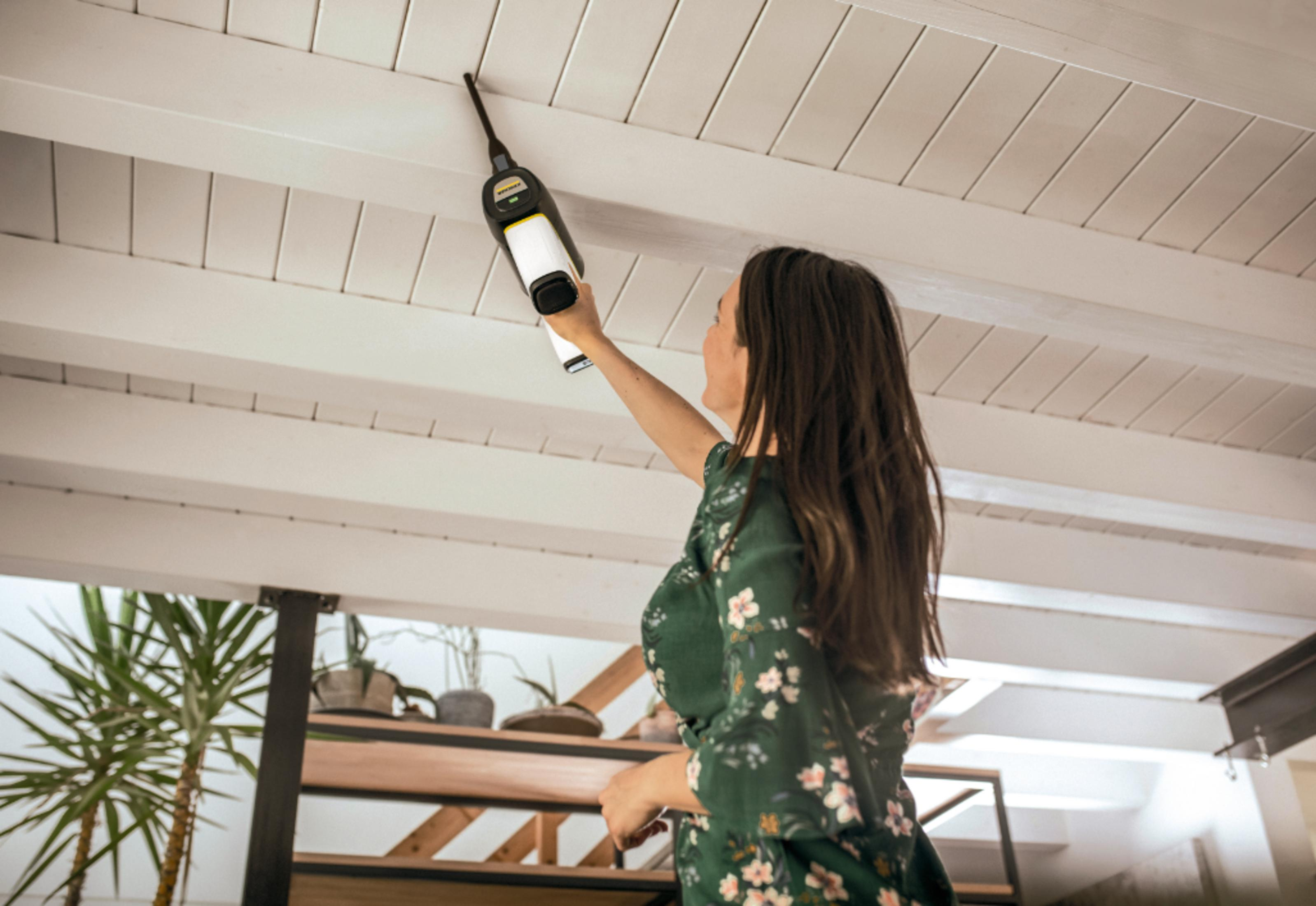 Kobieta w zielonej sukience czyści sufit odkurzaczem ręcznym.