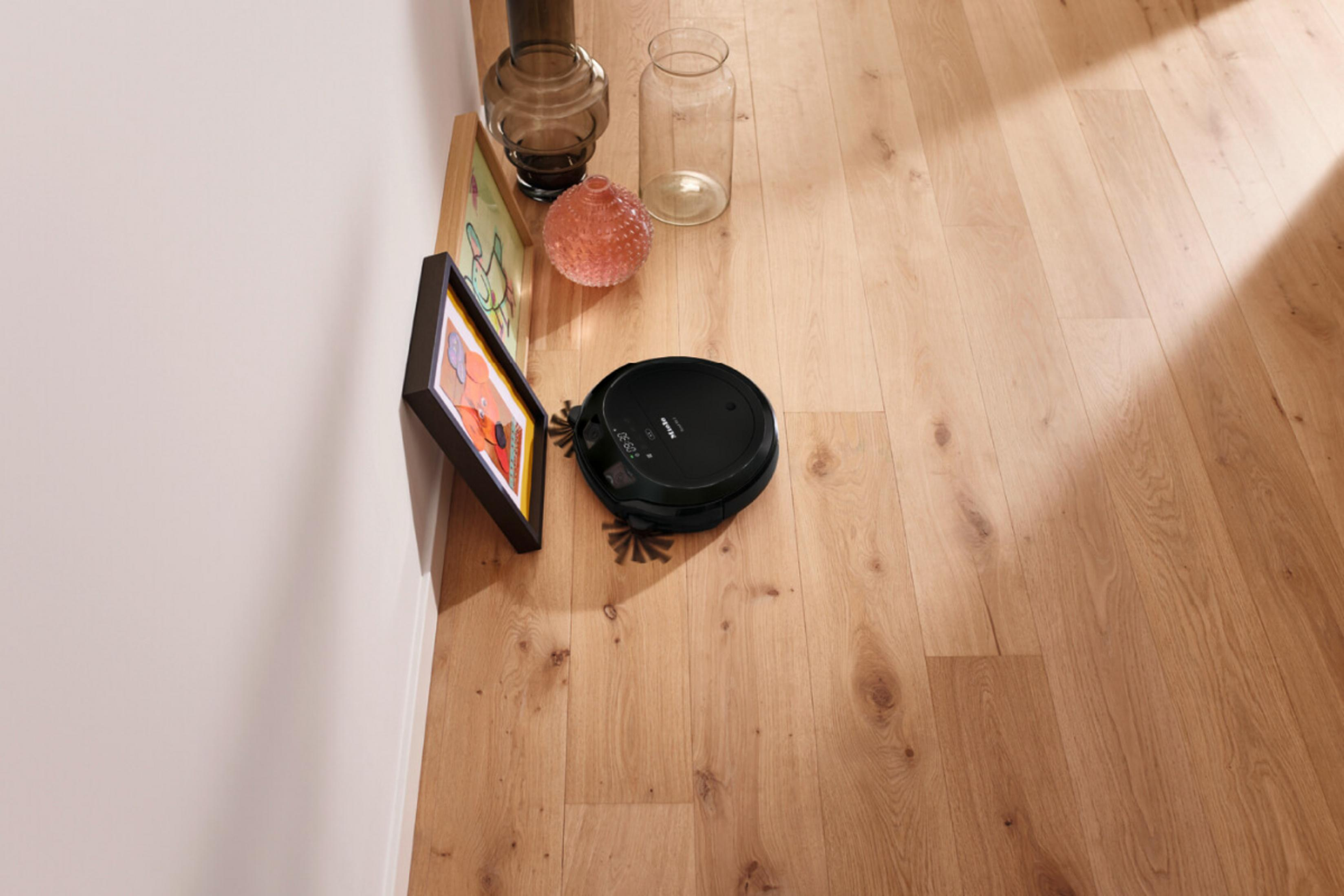 Ein schwarzer Saugroboter reinigt einen Holzboden neben einem gerahmten Bild und Vasen.