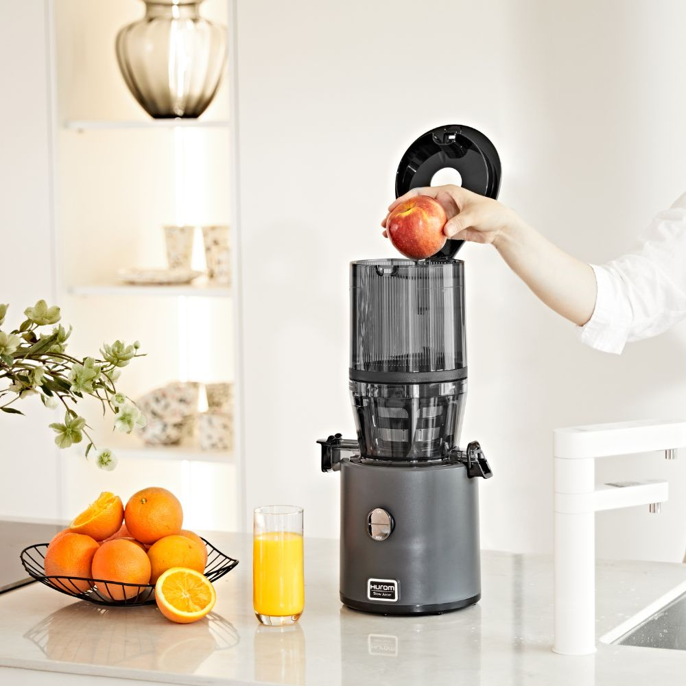 Eine Hand gibt einen Apfel in einen dunklen Entsafter neben Orangen und Saft.
