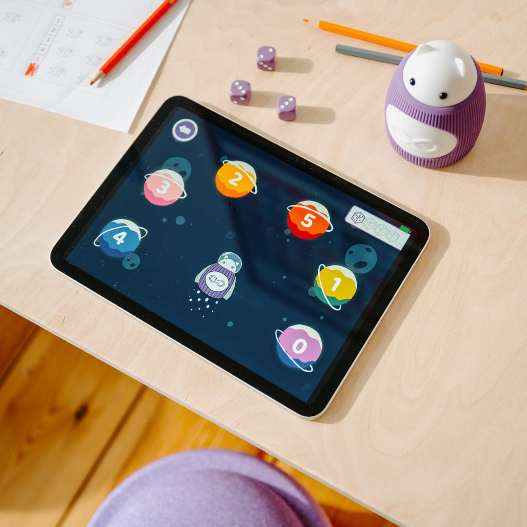 Ein Tablet mit einem Lernspiel, Würfel, Stifte und ein Spielzeug-Roboter liegen auf einem Holztisch.