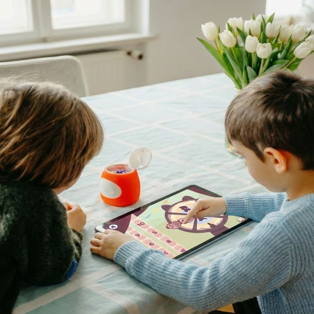 Zwei Kinder spielen ein Spiel auf einem Tablet. Ein orangefarbener Behälter steht daneben.