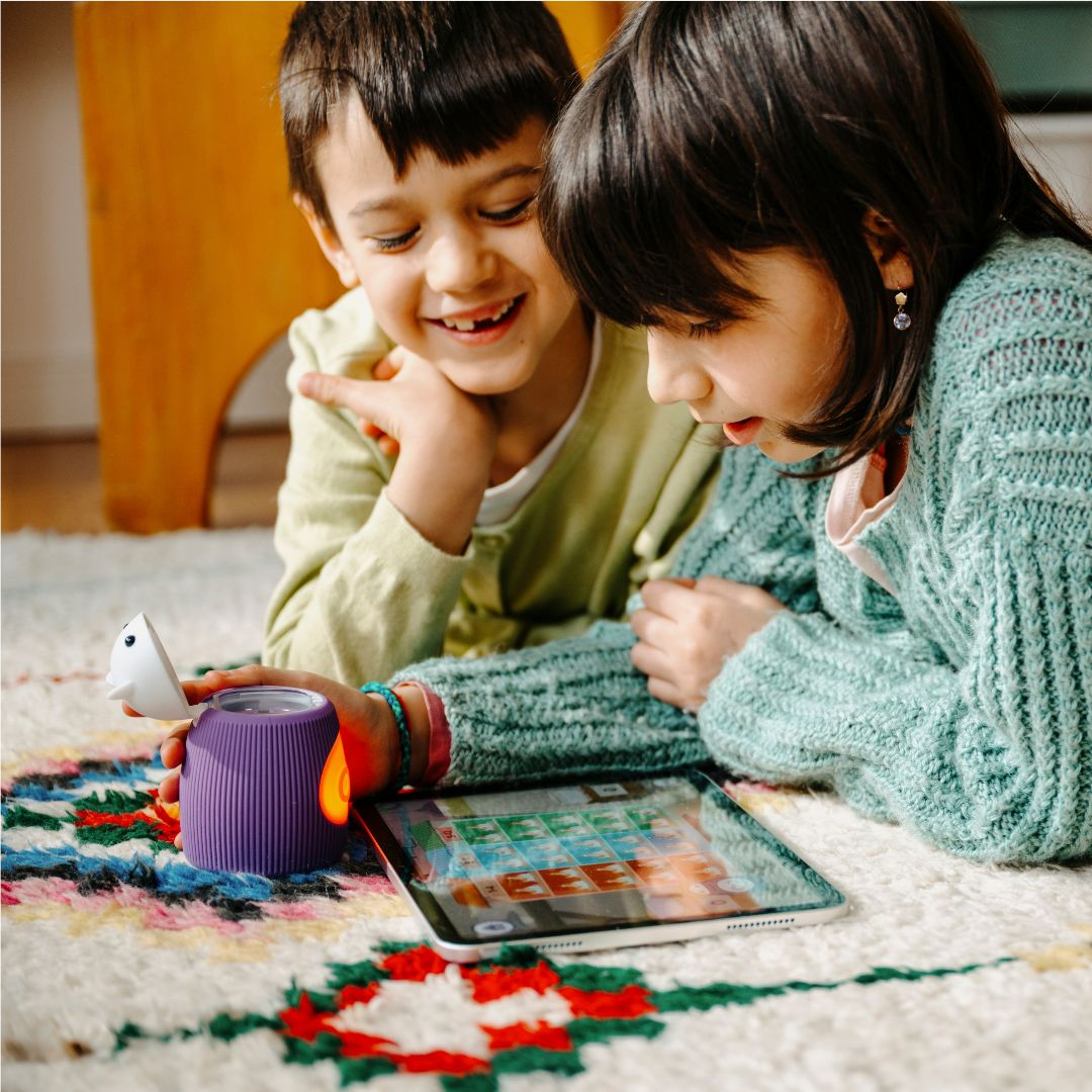 Zwei Kinder liegen auf einem Teppich und spielen mit einem Tablet und einem lila Spielzeug.