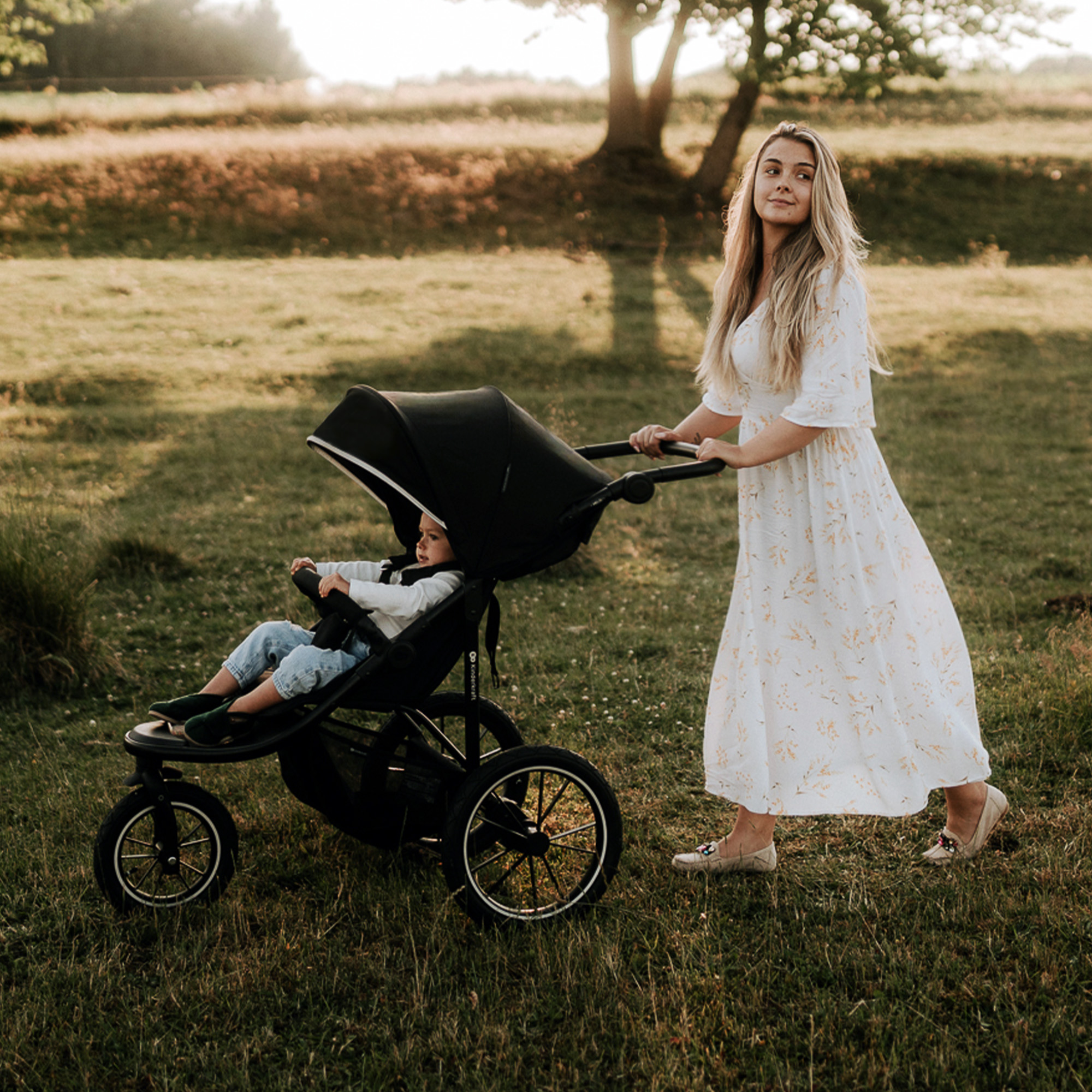Frau schiebt Kinderwagen mit Kind im Freien bei Sonnenuntergang im Gras.