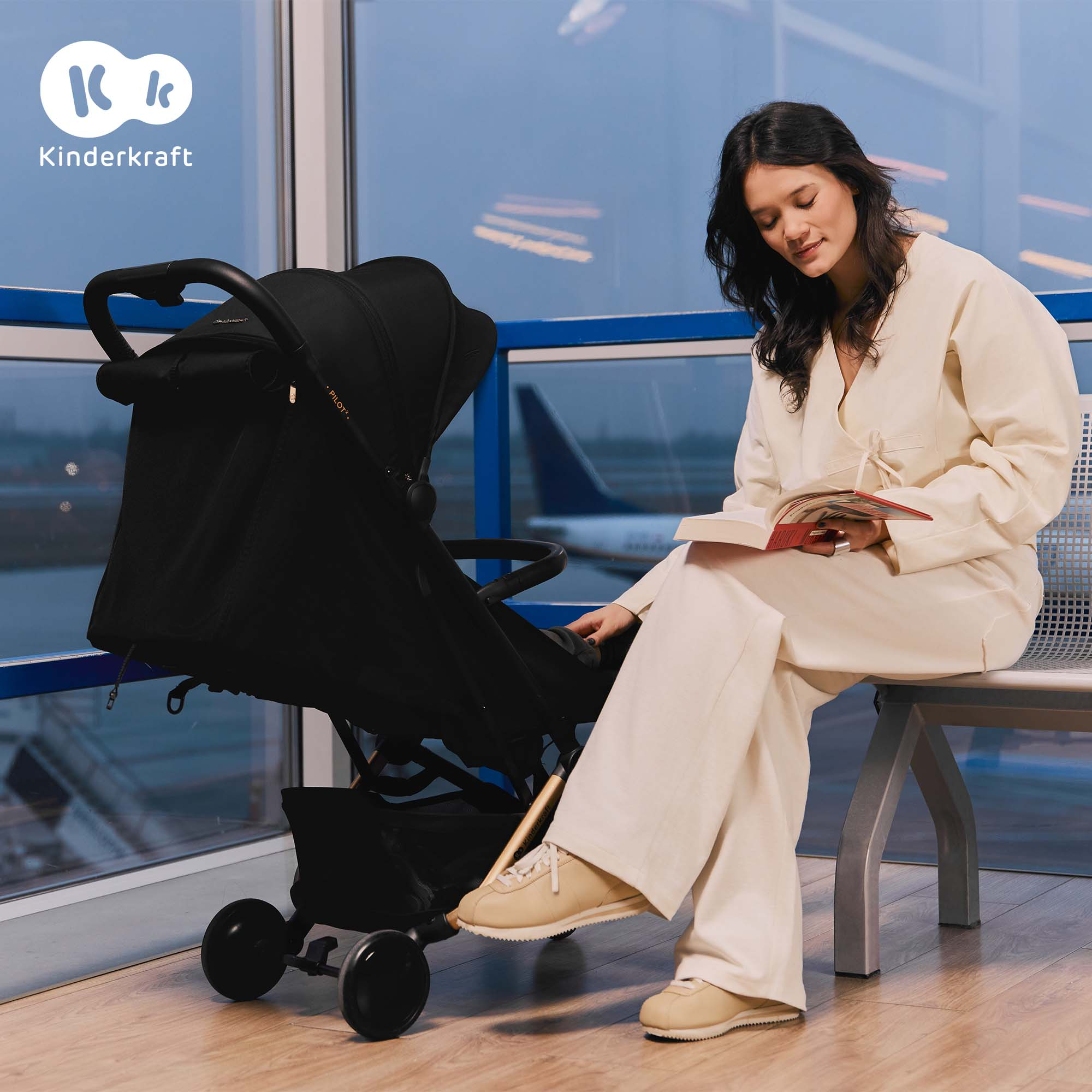 Frau liest Buch neben Kinderwagen am Flughafen.