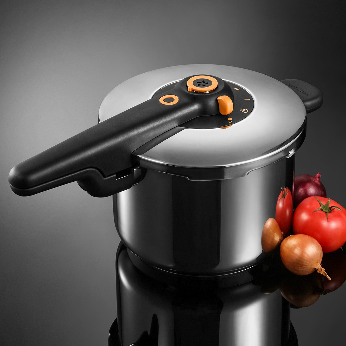 Edelstahl-Schnellkochtopf mit schwarzem Griff, daneben Zwiebeln und Tomaten.