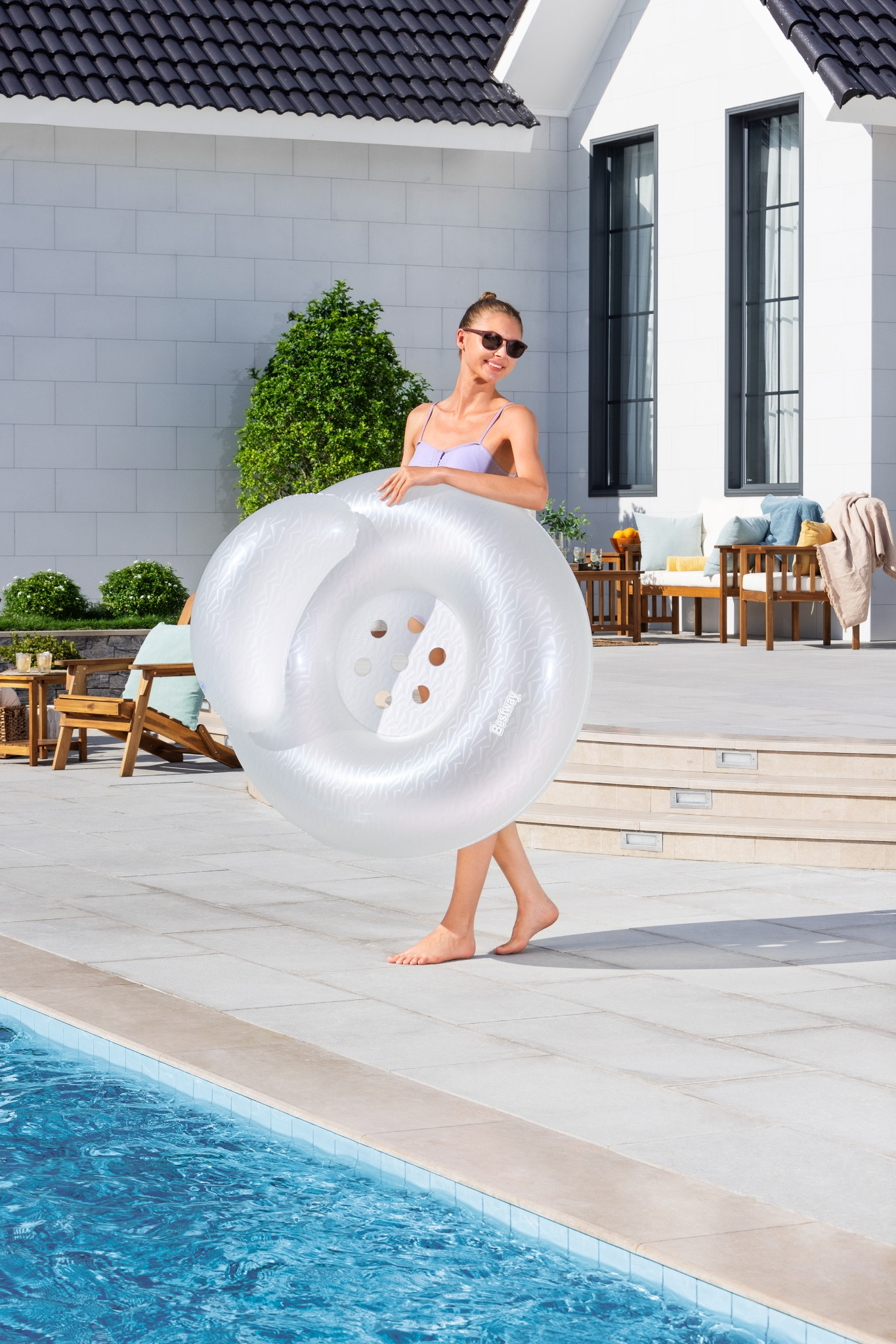 Frau in lila Bikini hält aufblasbaren Ring neben Pool auf gefliester Terrasse.
