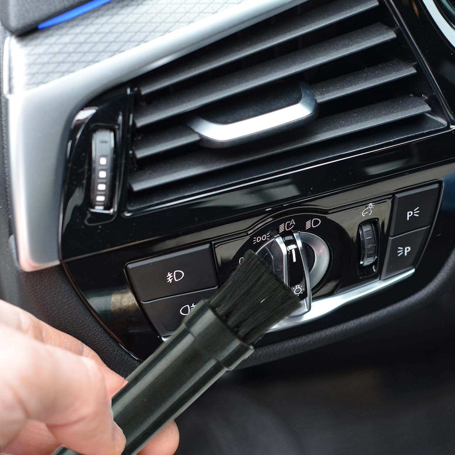 Eine Hand hält eine schwarze Bürste in der Nähe eines Auto-Lüftungsschlitzes und Bedienelements.