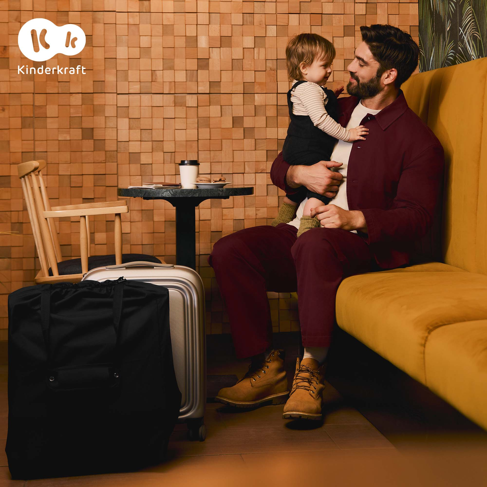 Mann mit Baby auf gelbem Sofa, Gepäck daneben. Holzwand im Hintergrund.