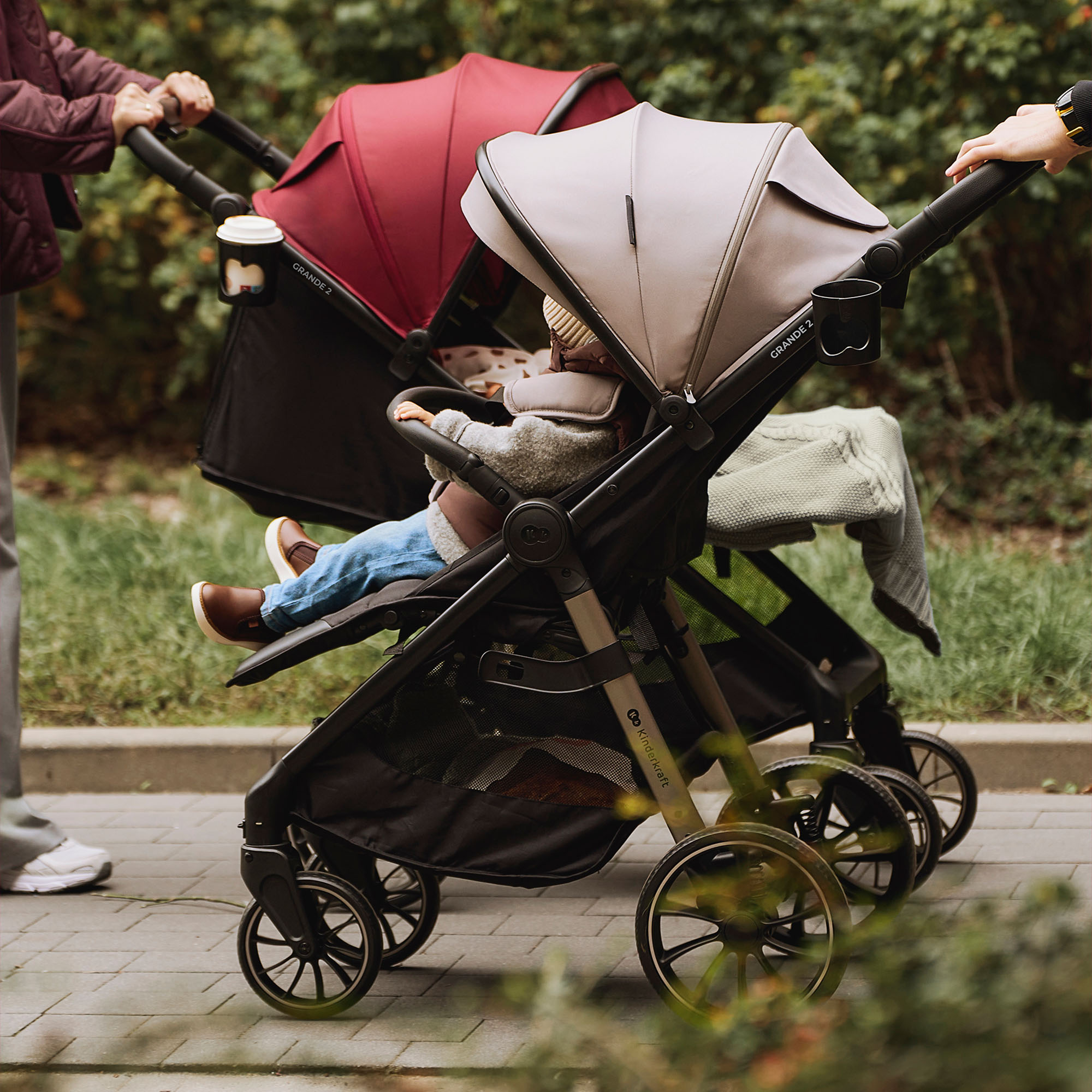 Zwei Kinderwagen nebeneinander, einer mit rotem Verdeck, einer mit beige. Ein Baby sitzt im vorderen Wagen.