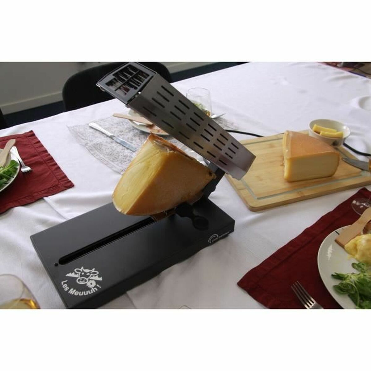 Raclette-Grill mit Käse, Brot und Salat auf einem Tisch mit weißer Decke.