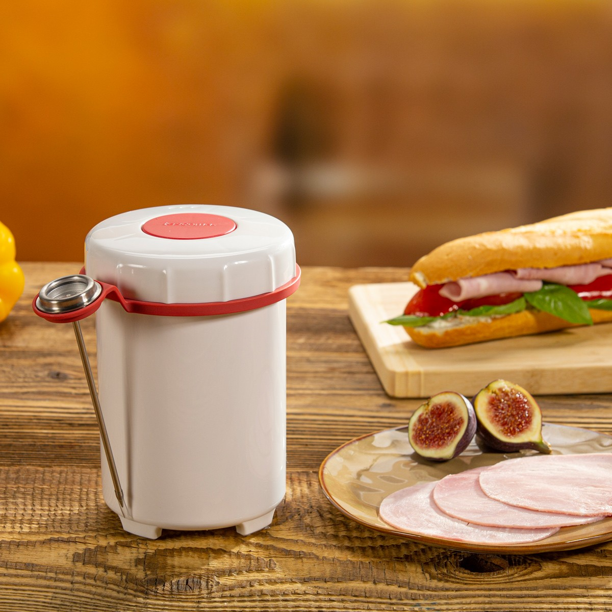 Ein weißer Behälter mit rotem Deckel und Thermometer neben Sandwich und Schinken.