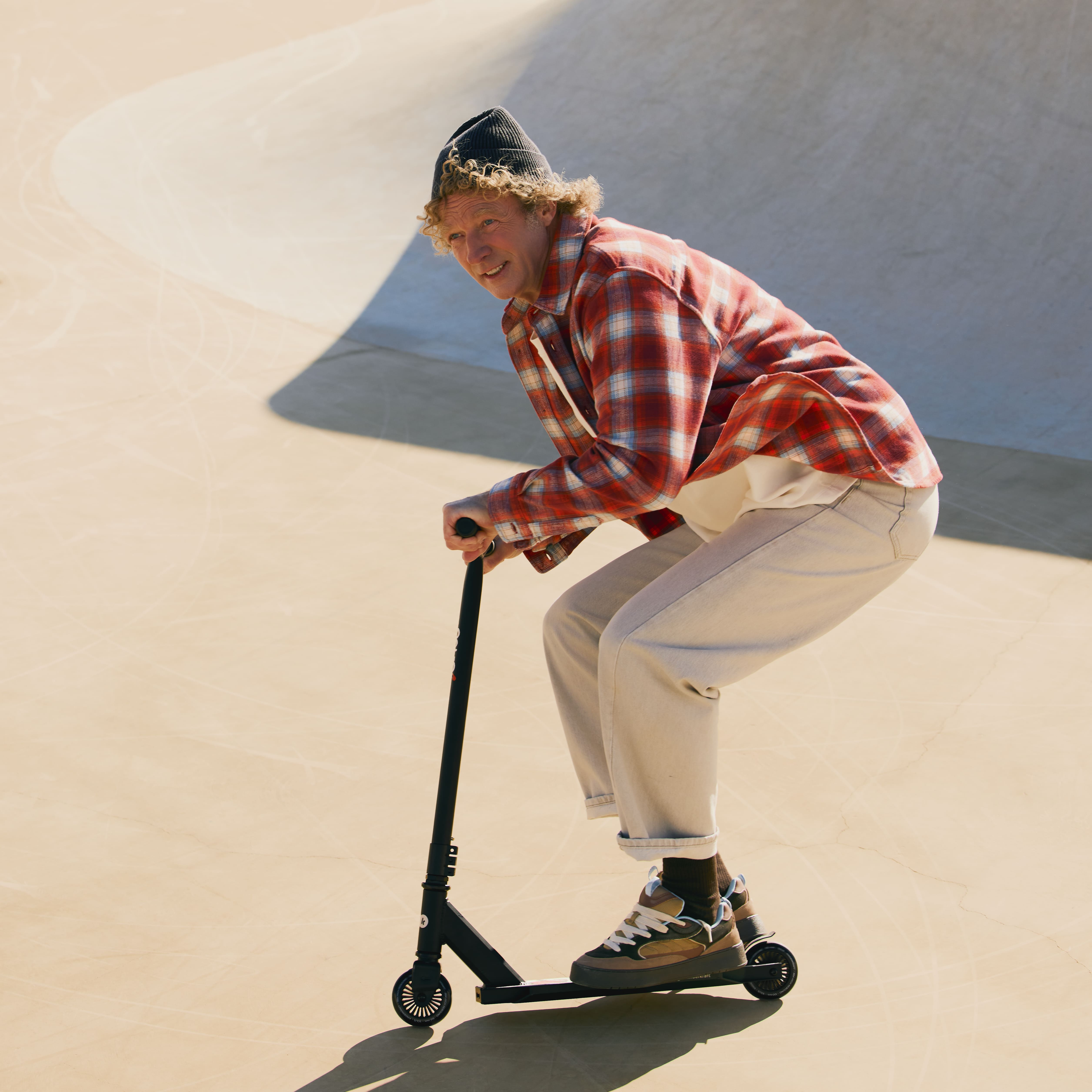 Ein Mann mit lockigem Haar fährt einen Roller in einem Skatepark.