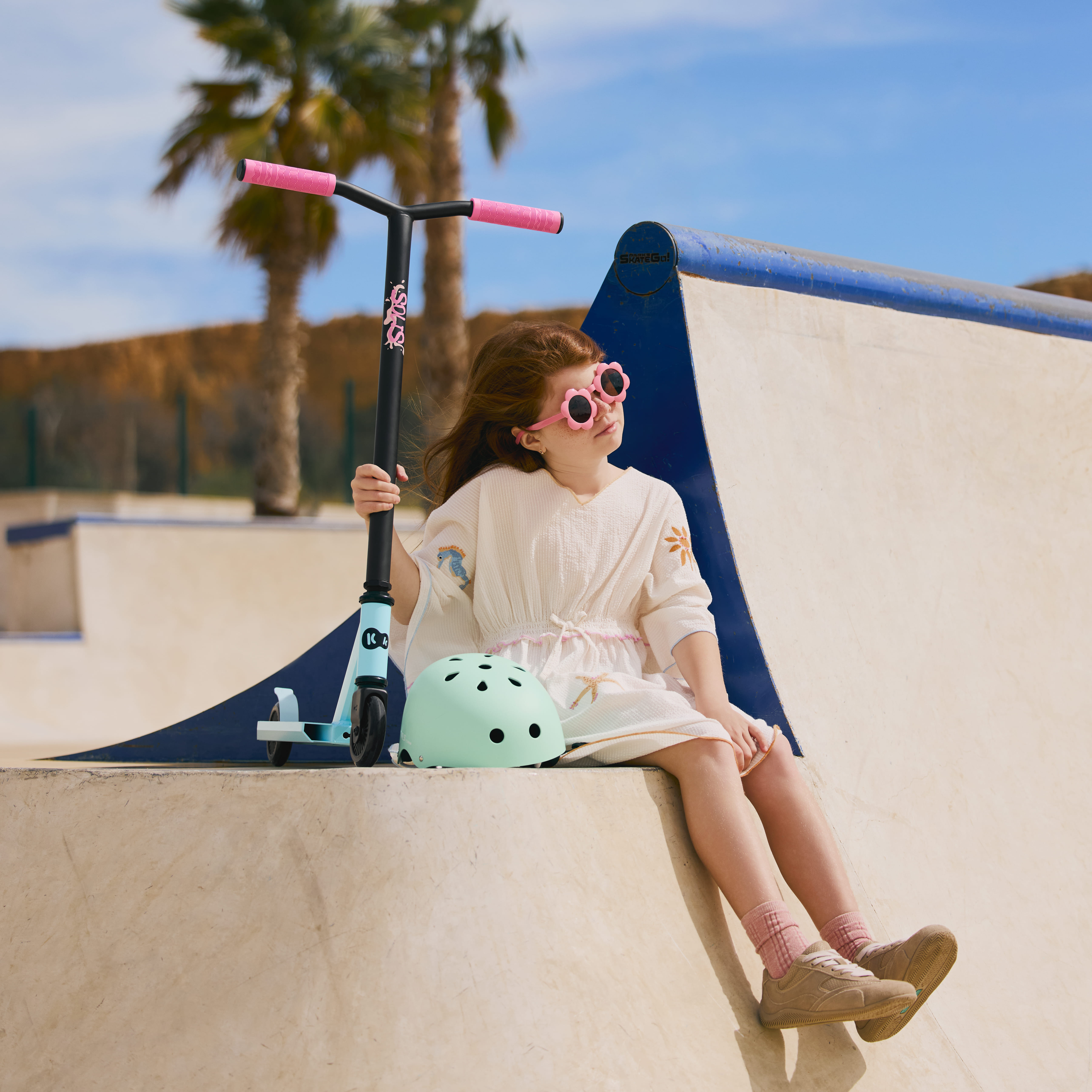 Mädchen mit Roller und Helm sitzt auf Rampe, Palmen und blauer Himmel im Hintergrund.