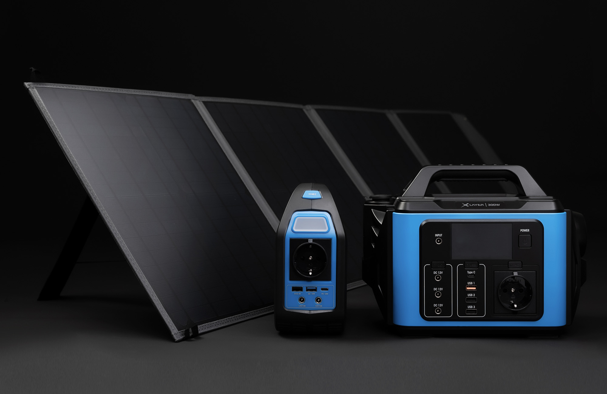 Ein faltbares Solarpanel steht hinter zwei blauen und schwarzen Powerstations auf dunklem Grund.