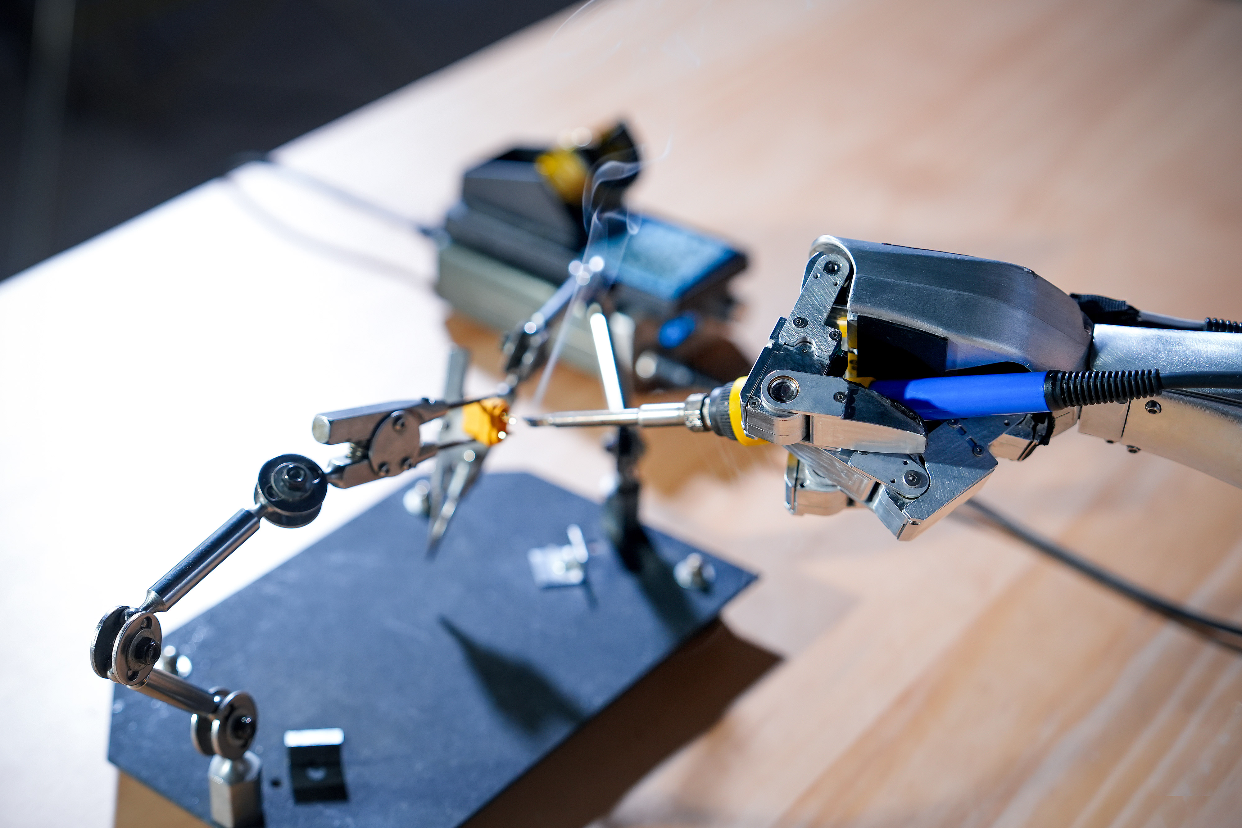 Ein Roboterarm hält eine Lötpistole über ein goldenes Objekt, Rauch steigt auf.